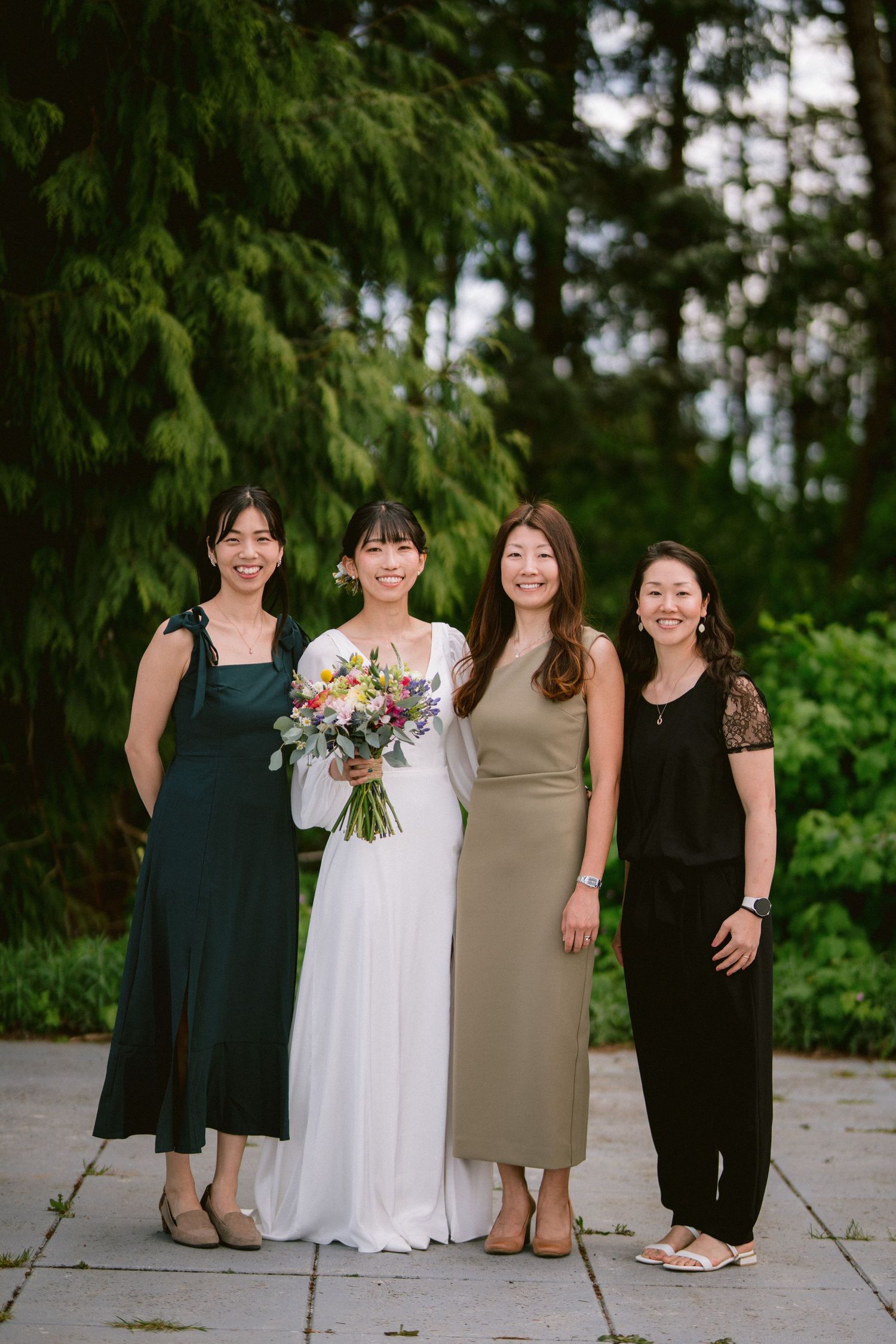 Four women in elegant dresses pose together at a wedding, with the bride holding a colorful bouquet.