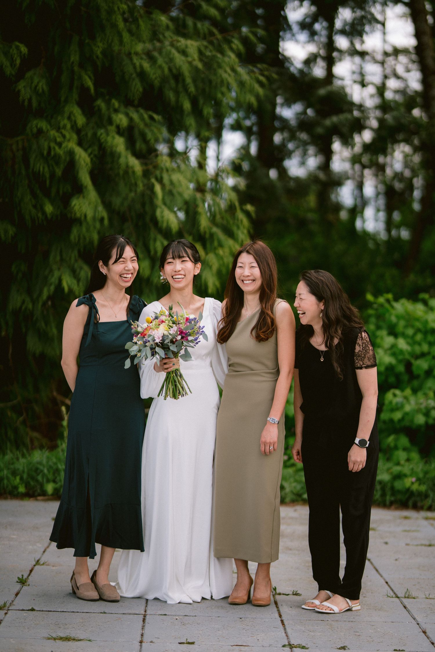A bridal party in elegant dresses poses together against a backdrop of evergreen trees.