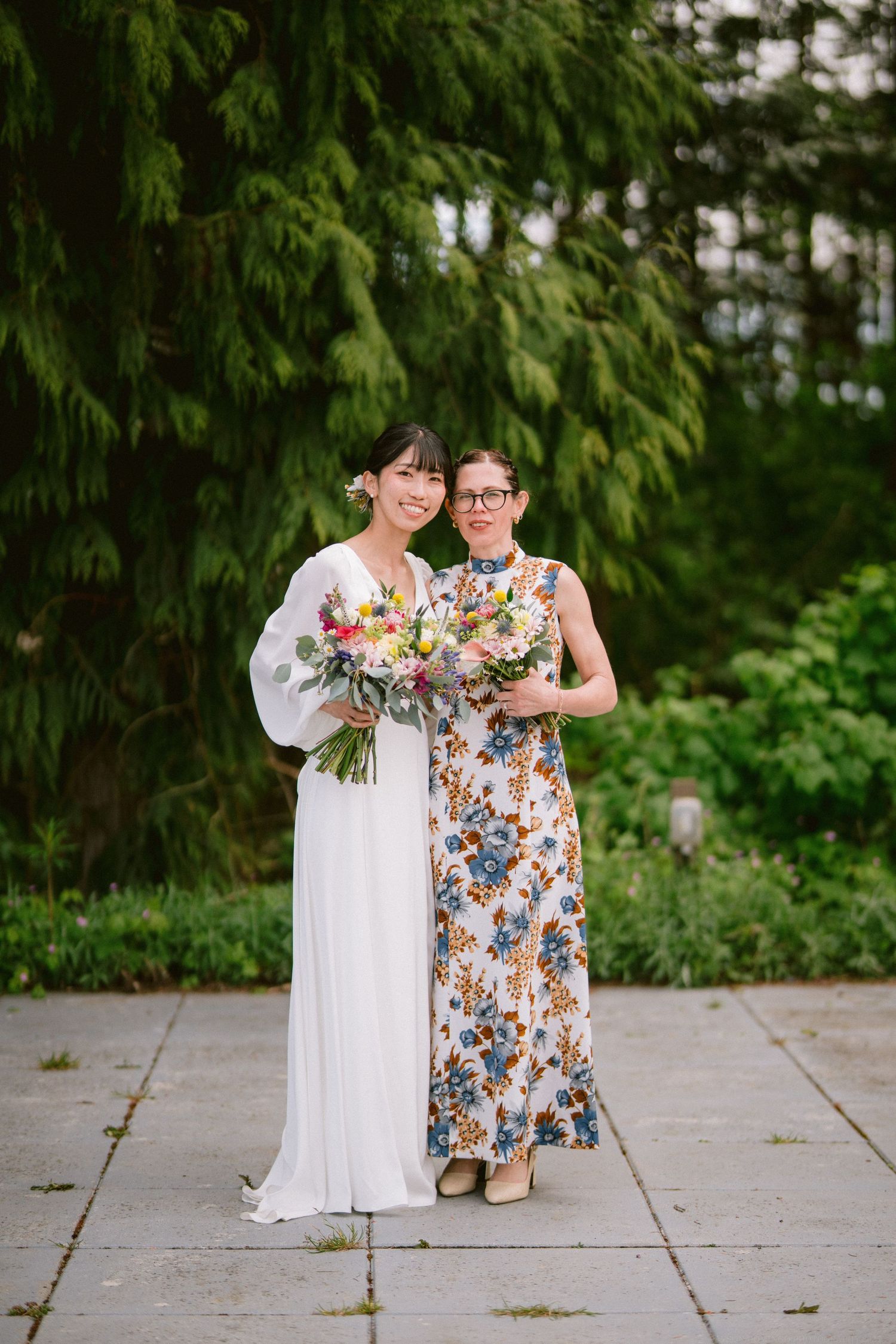 Two guests hold matching floral bouquets while posing on a stone pathway surrounded by evergreens.