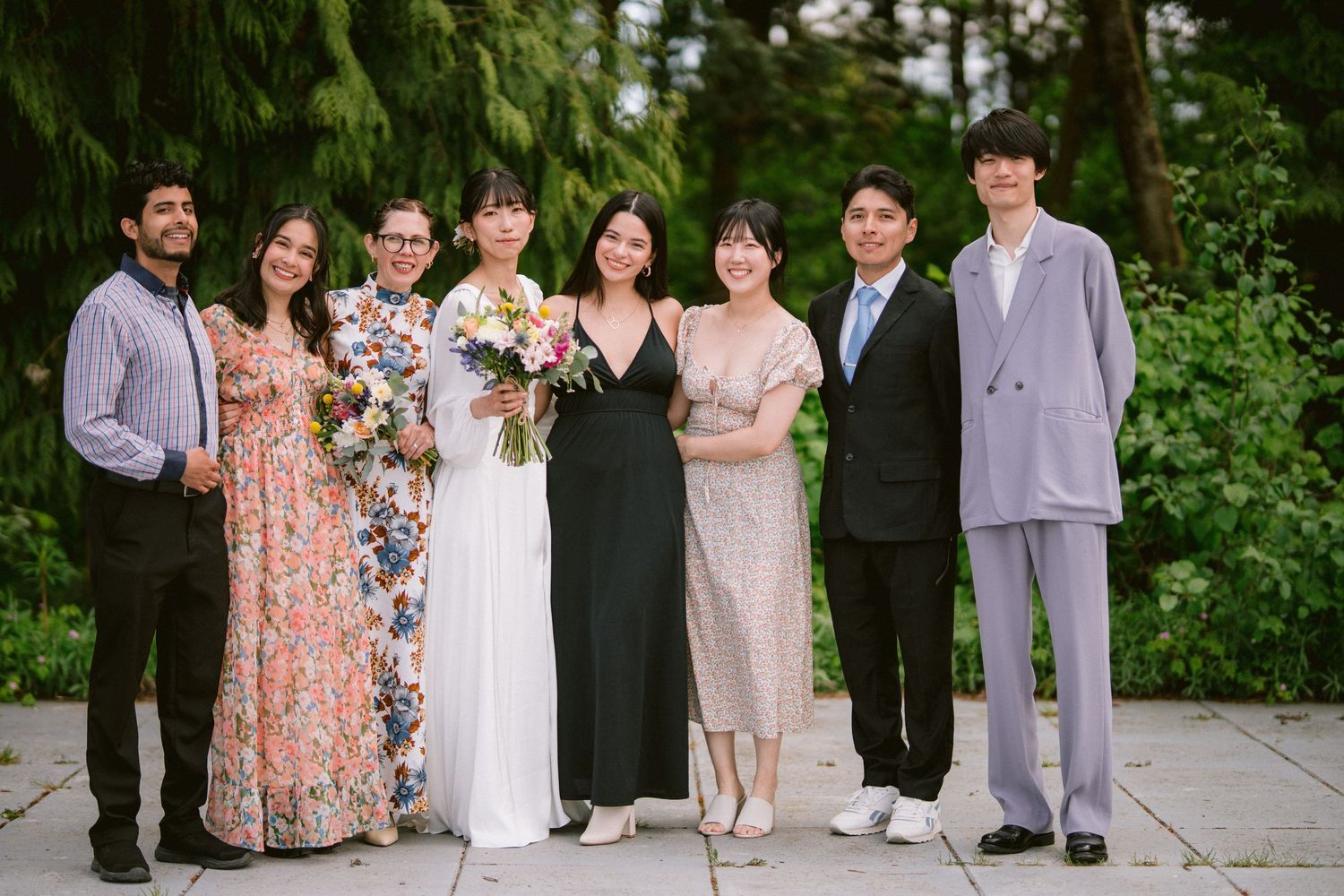 A formal group in elegant attire poses together outdoors against a backdrop of green trees and foliage.