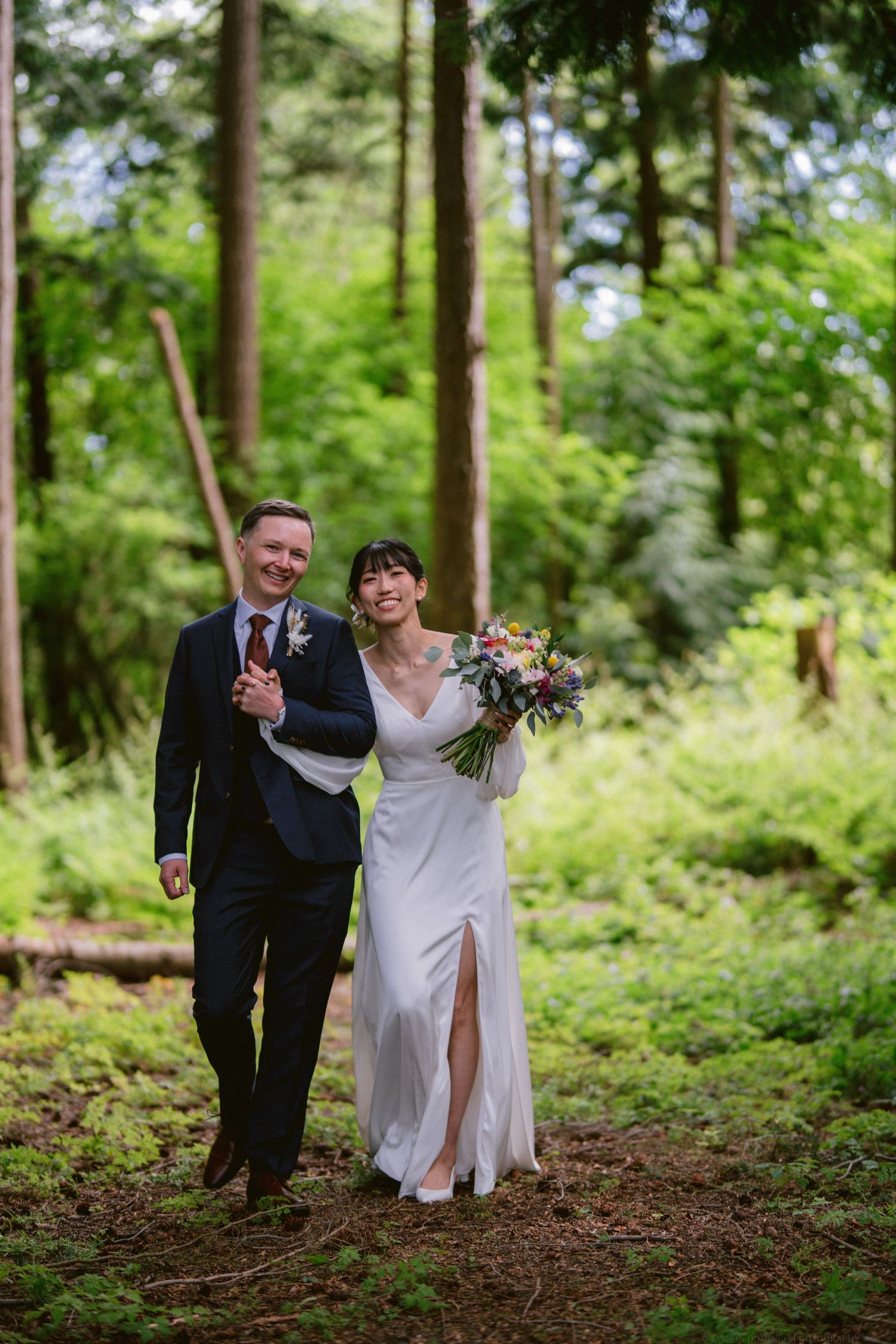 A wedding couple walks together on a forest path while carrying a colorful bouquet of flowers.