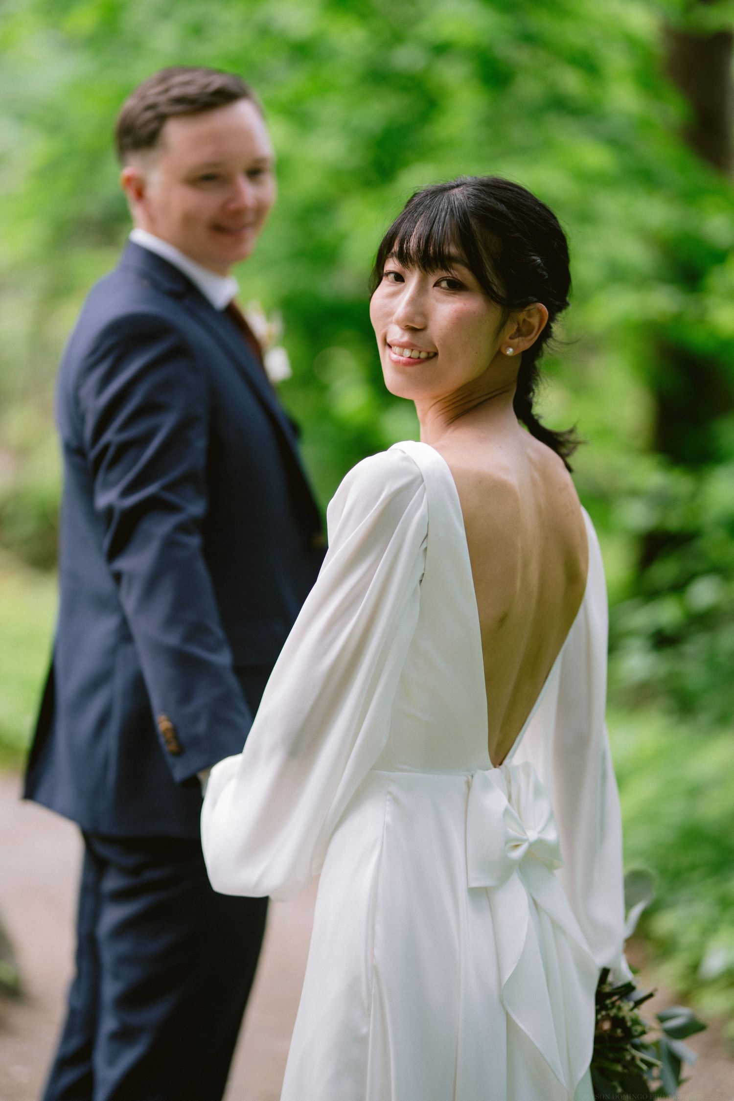A bride in an elegant white dress with an open back poses outdoors in a garden setting.