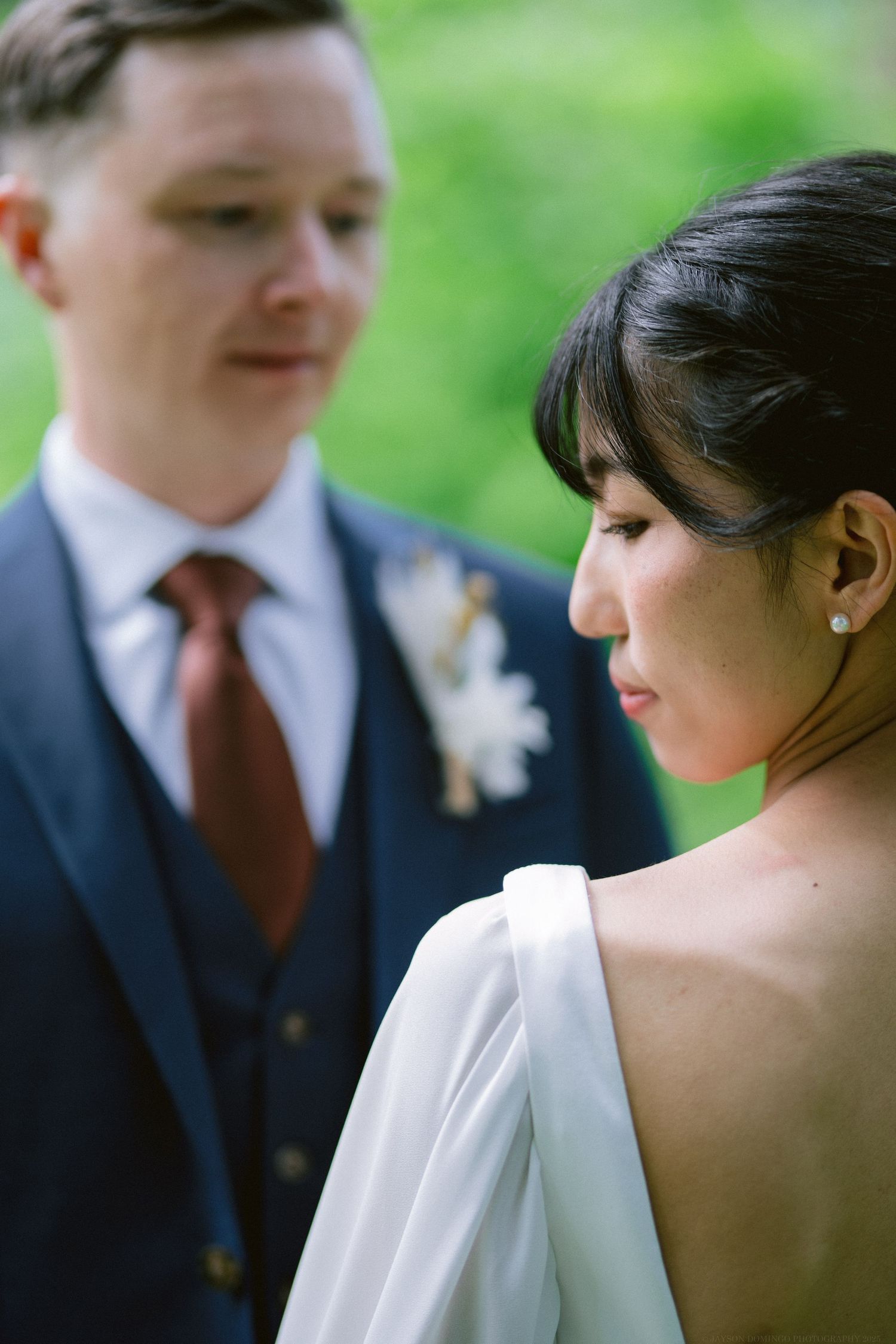 A close-up shot of a couple in wedding attire standing together against a green garden backdrop.
