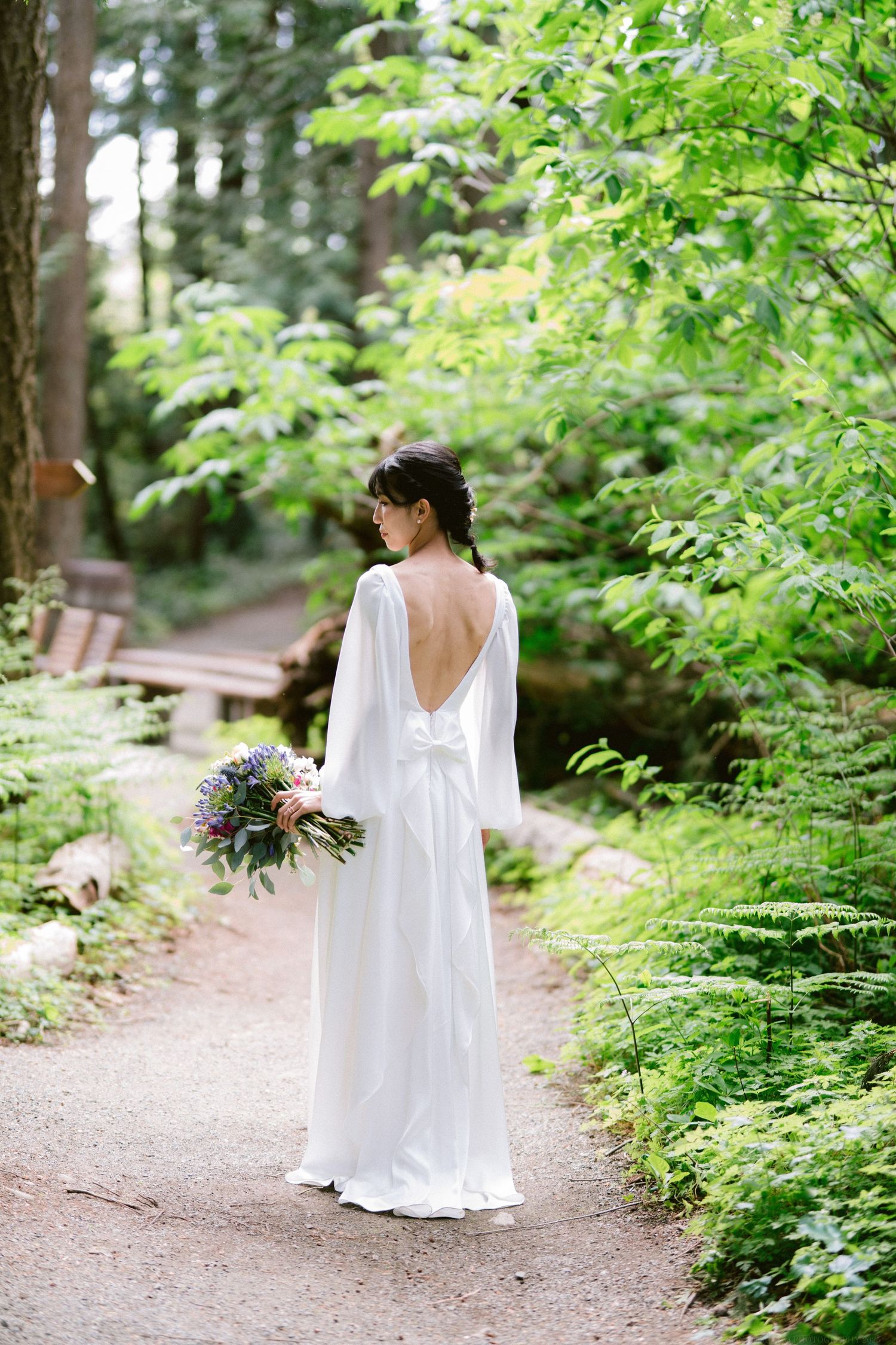 Someone in a flowing white wedding dress with an open back holds a bouquet while standing on a forest path surrounded by greenery.