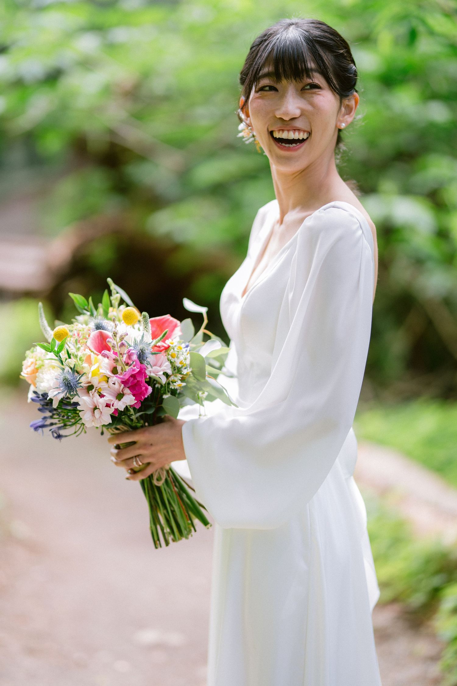 A joyful bride in a long-sleeved white dress laughs while holding a colorful floral bouquet in a garden.