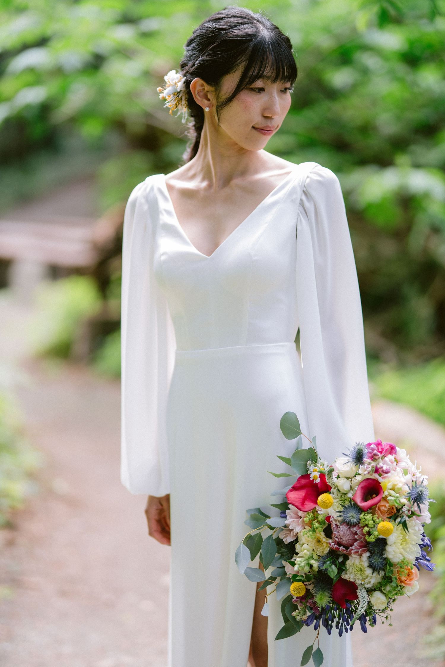 Bride in elegant white dress with long sleeves holds vibrant wildflower bouquet on forest path.