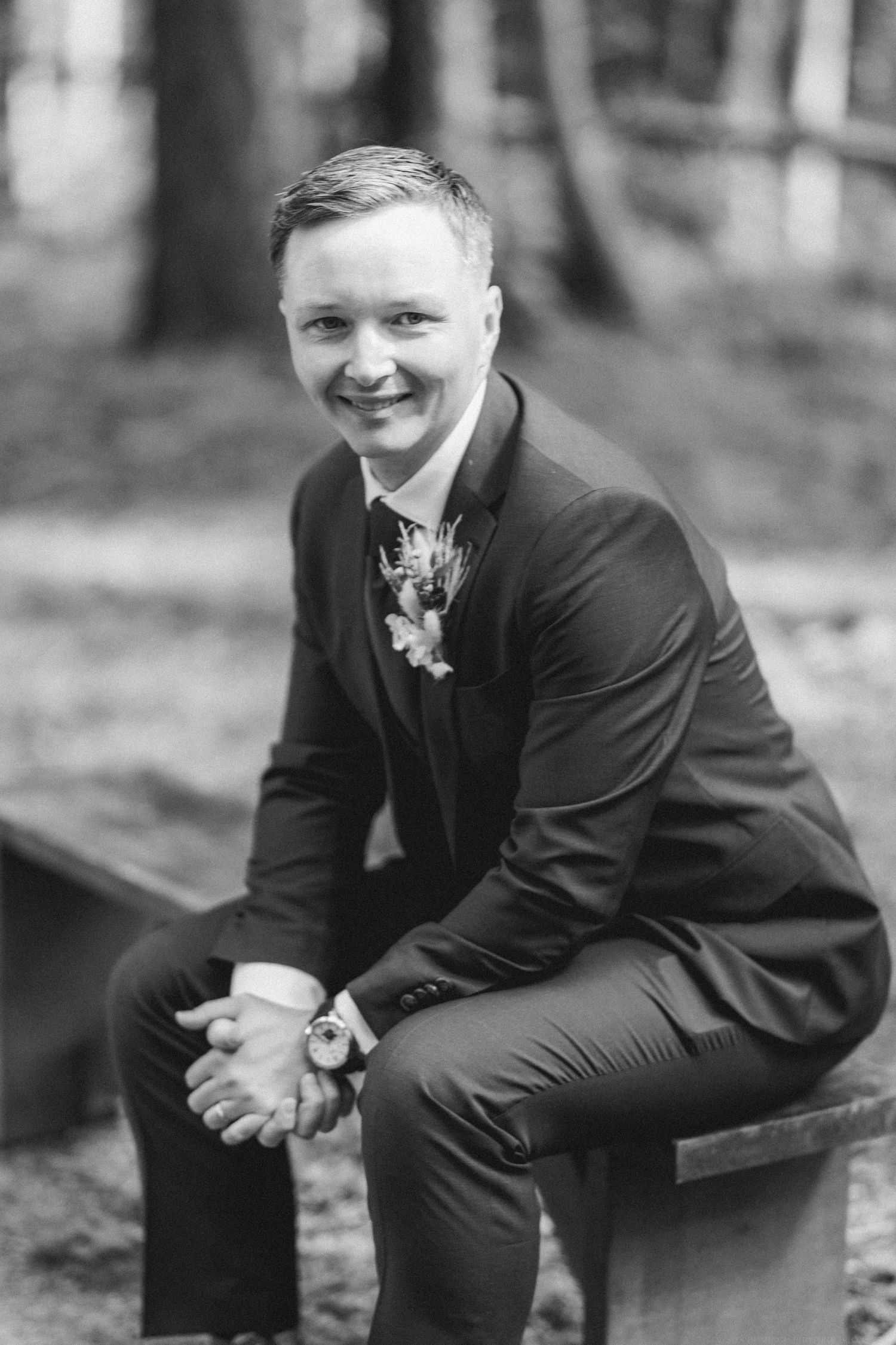 Black and white portrait of groom in formal suit sitting on bench in forest setting.
