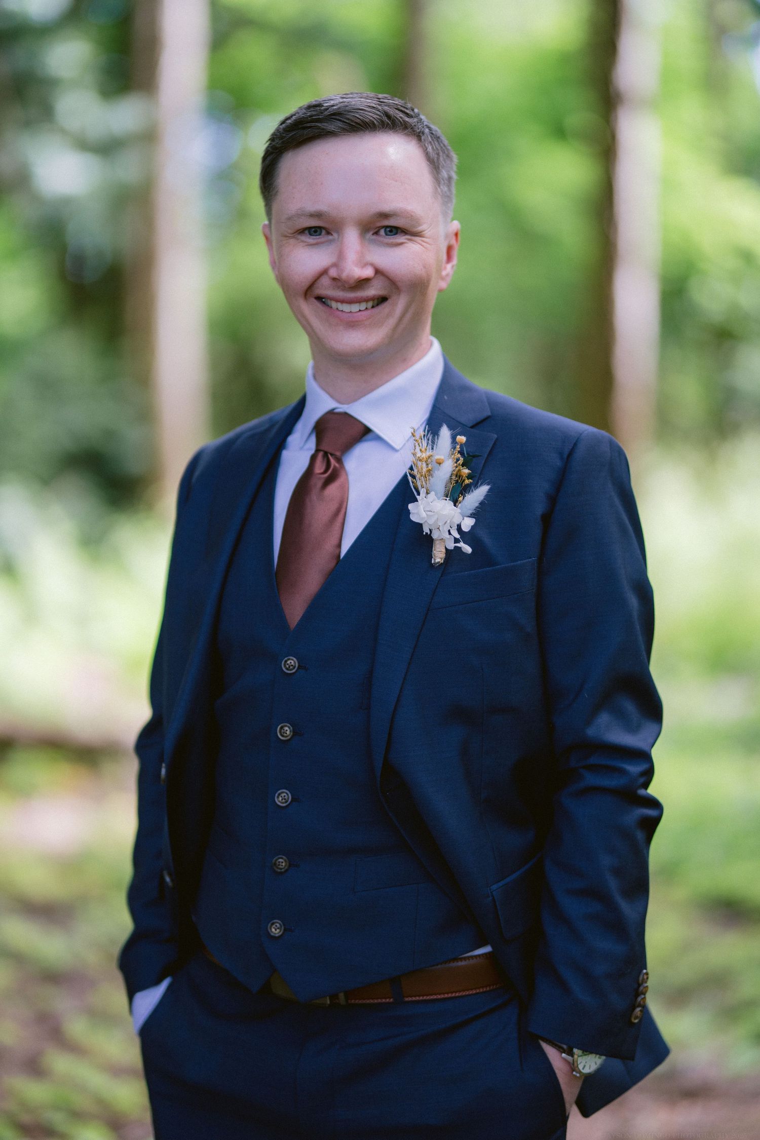 Groom in navy three-piece suit with burgundy tie and boutonniere smiles in outdoor forest setting.