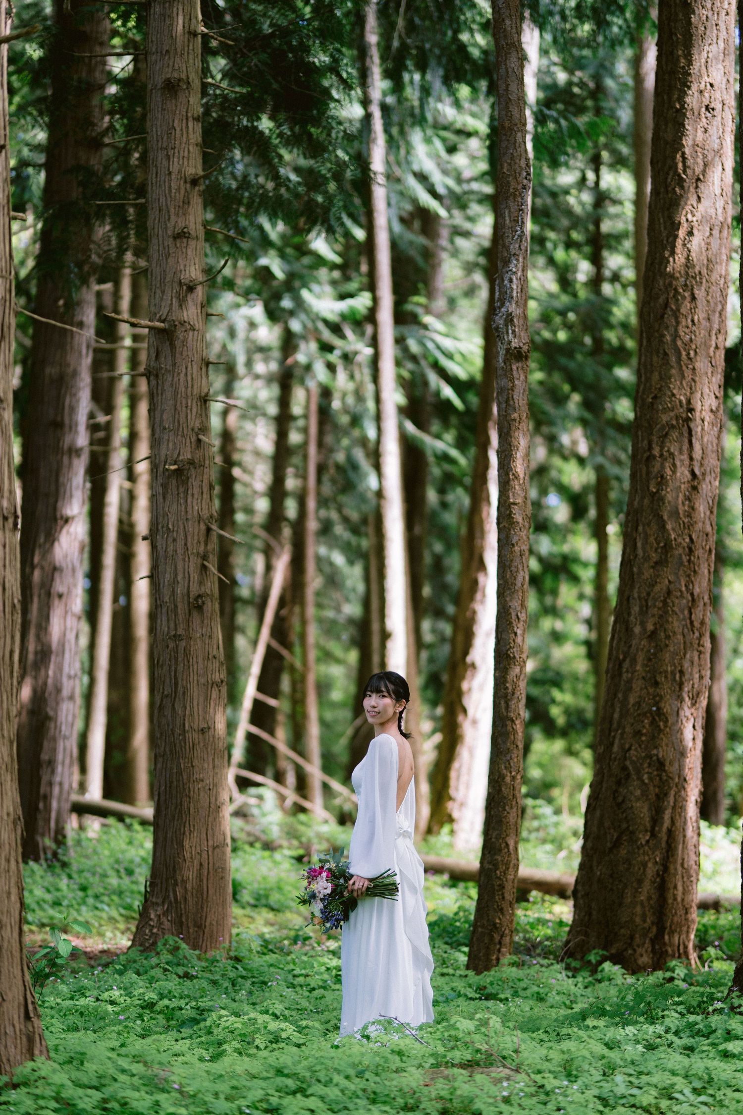 A bride in a white dress stands alone in a serene forest setting holding a bouquet.