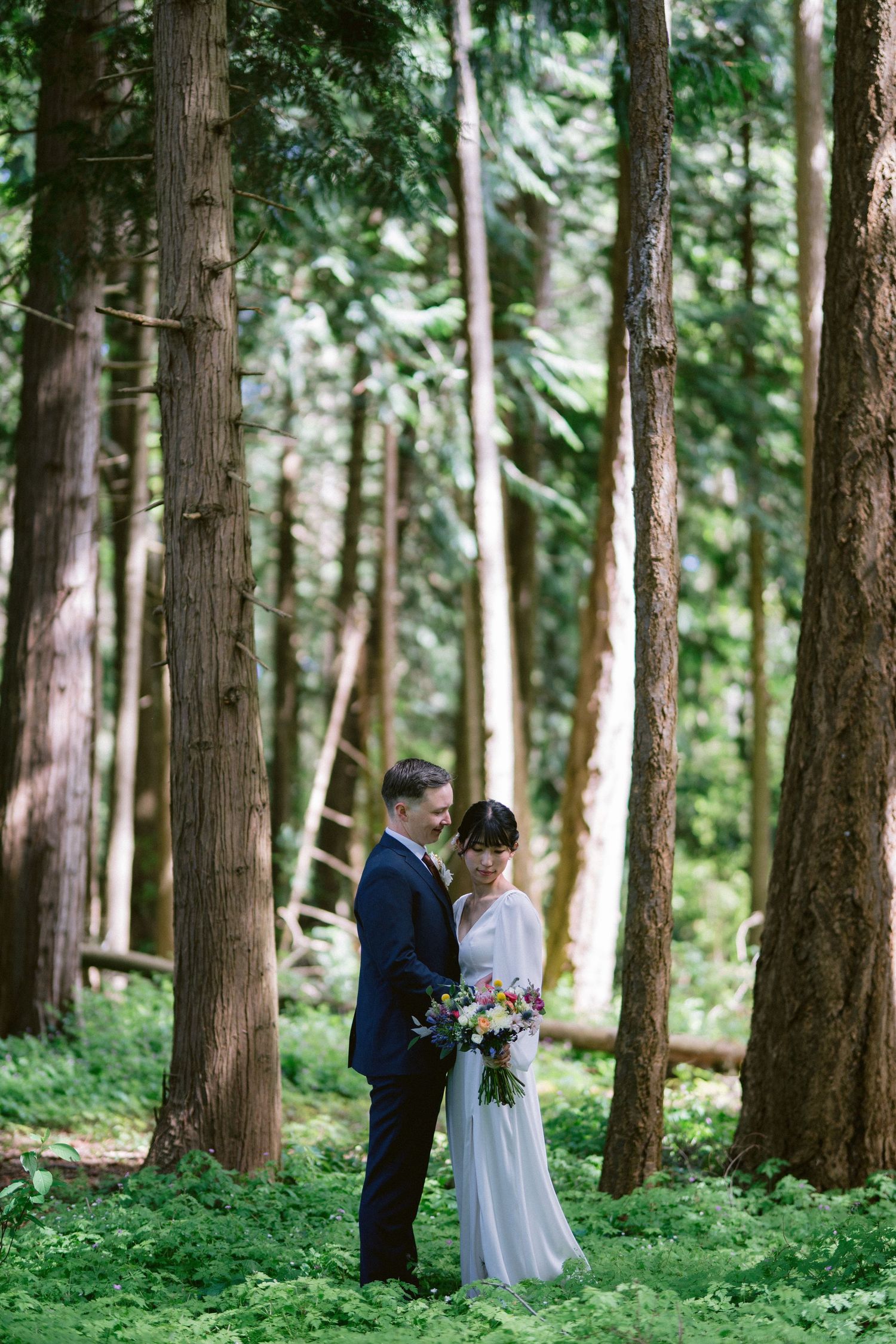 Newlyweds share an intimate moment among tall trees in a lush forest setting.