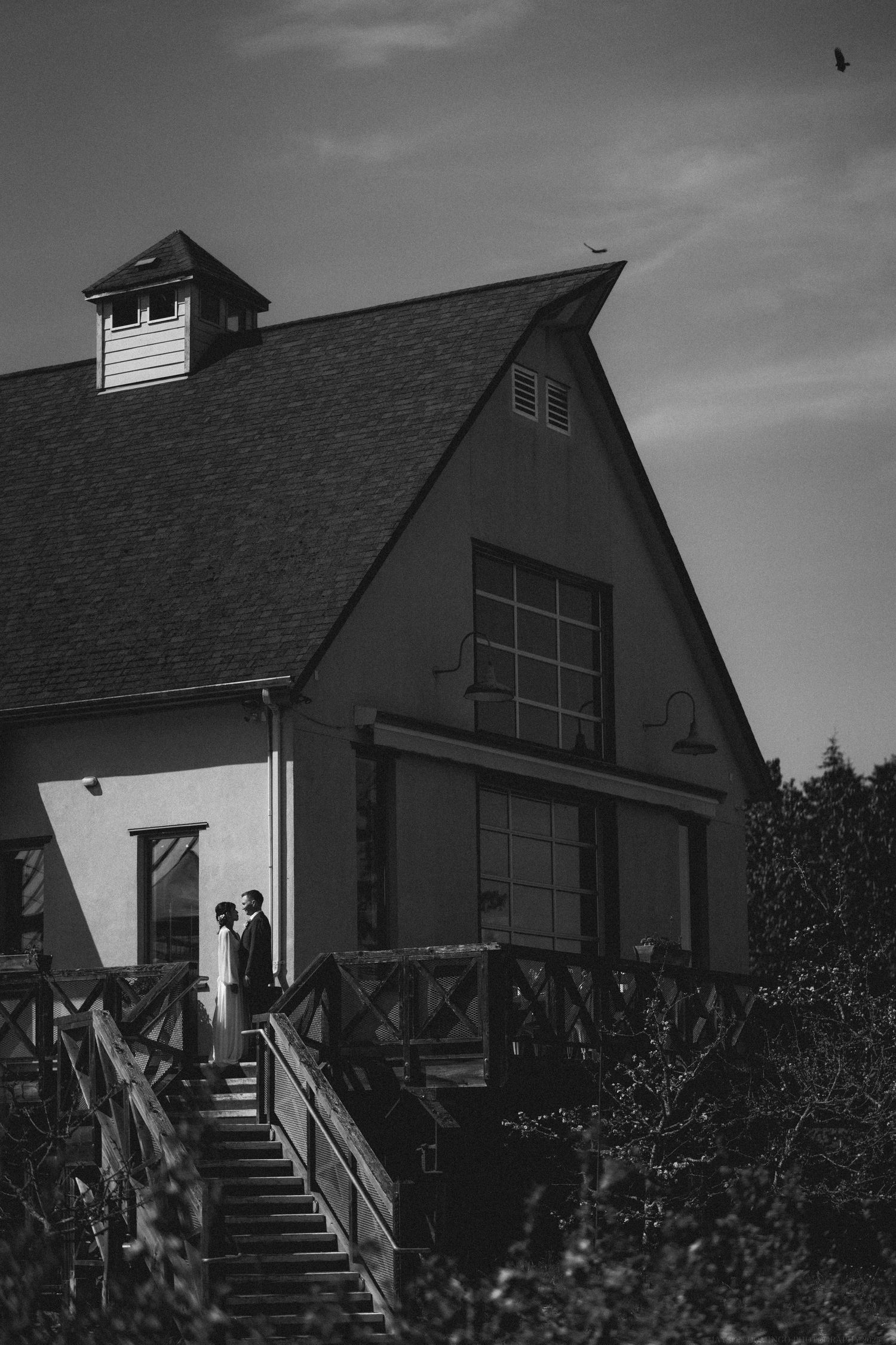A black and white photo of a rustic barn-style building with figures standing on the entrance stairs.