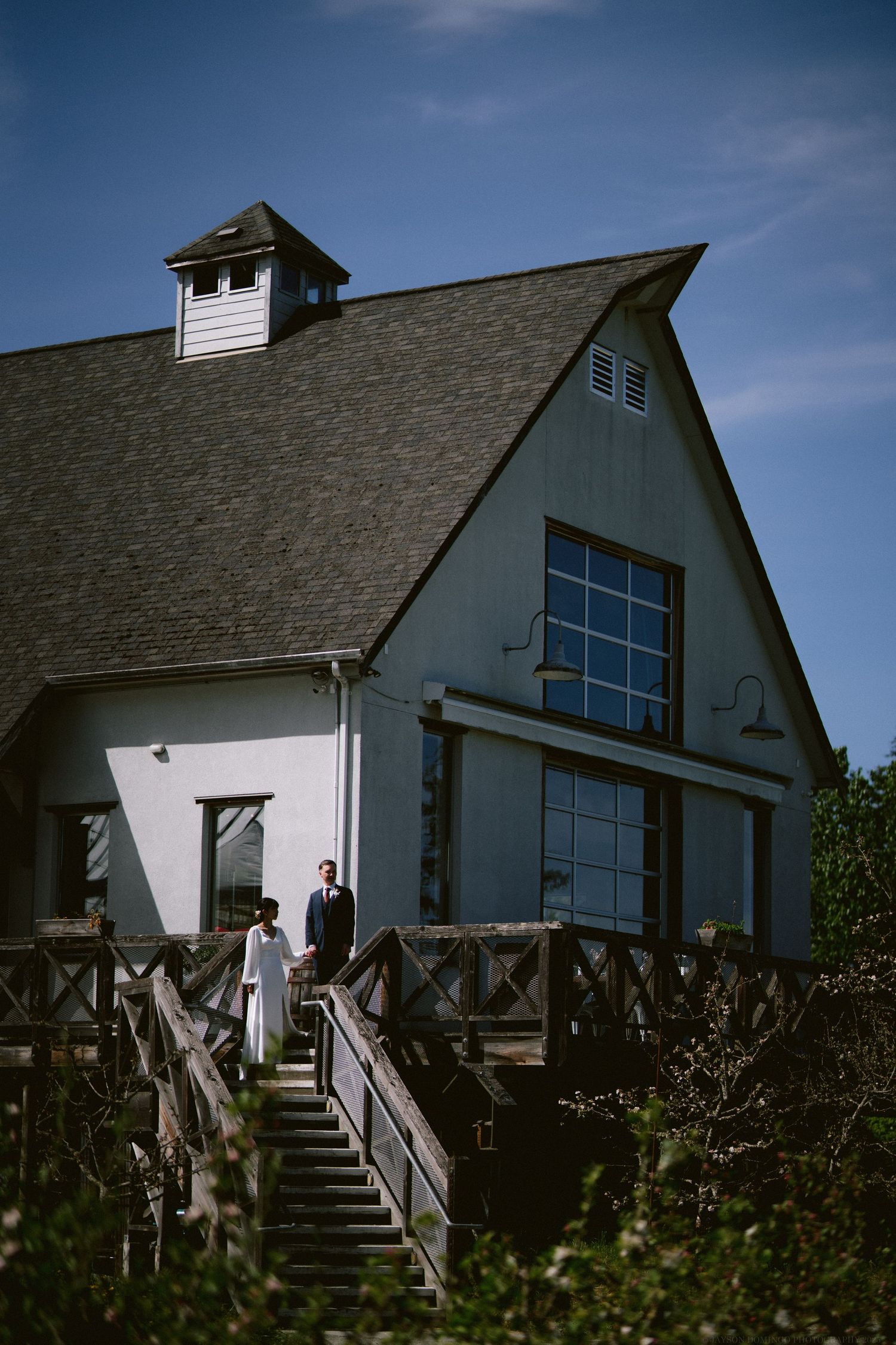 A couple stands on wooden steps leading to a white barn-style building with large windows under blue sky.