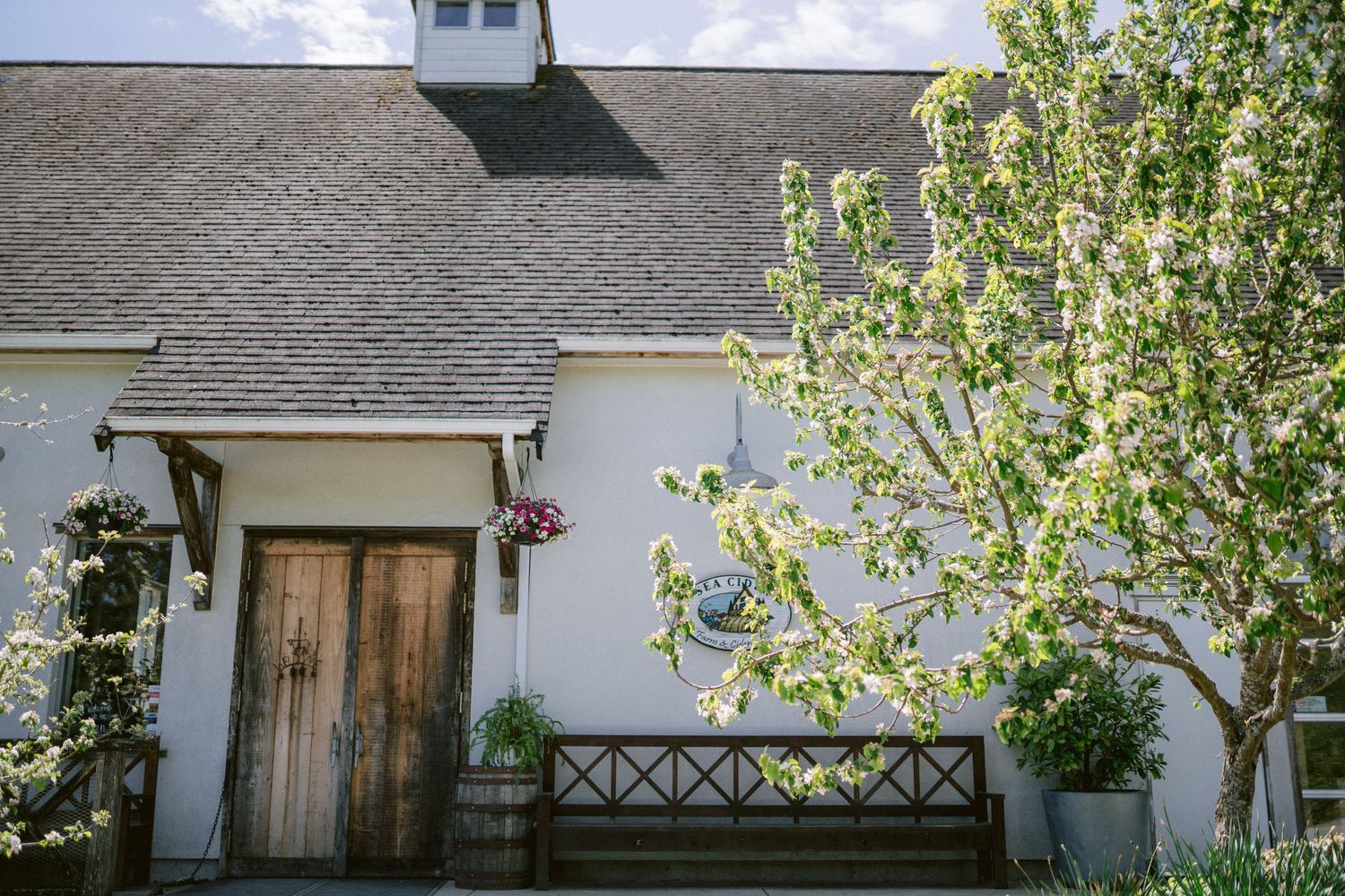 White church building exterior with wooden door, hanging flower baskets, and wooden bench against stucco wall.