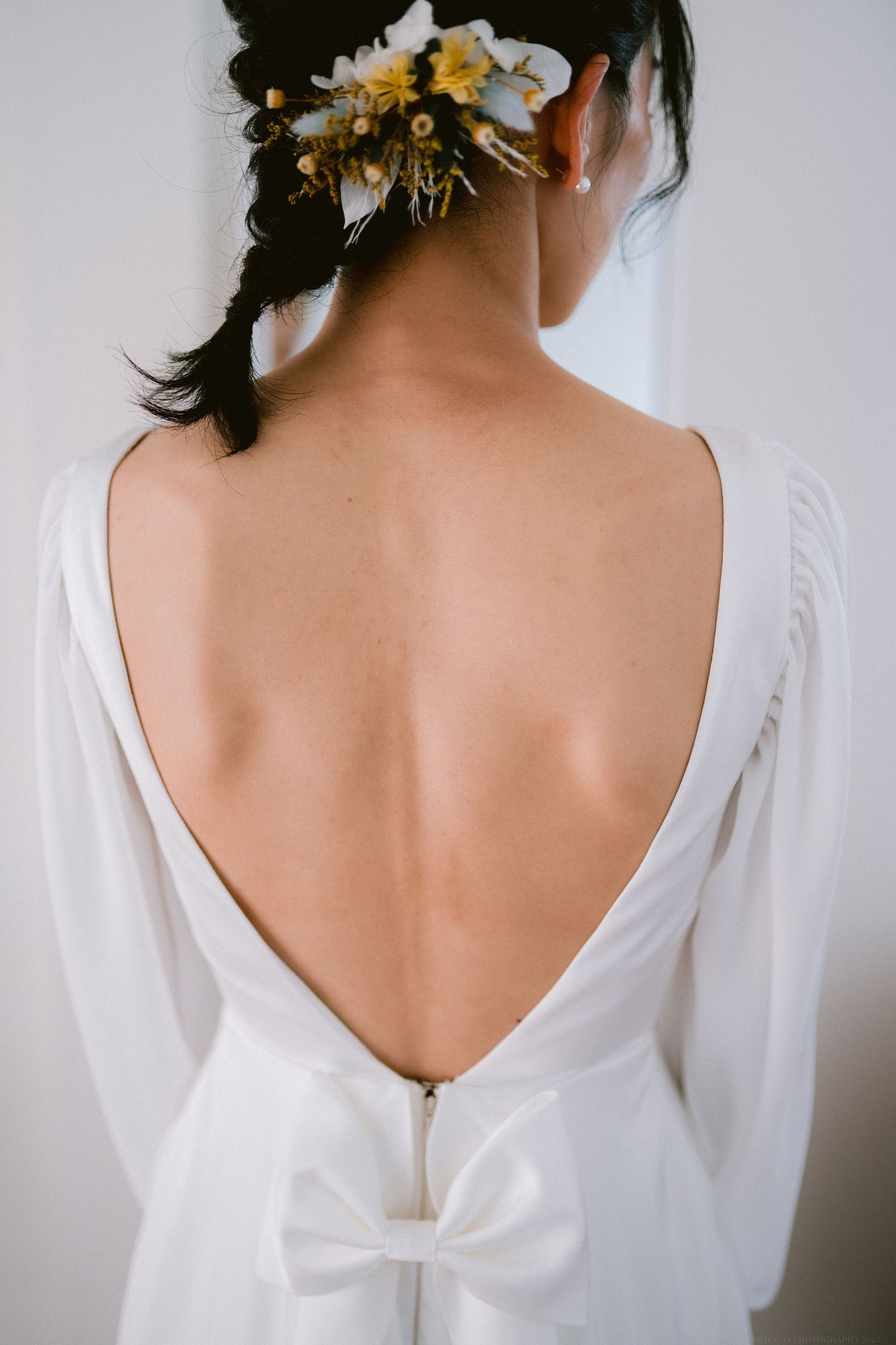 Back view of bridal gown showing plunging V-back with bow detail and floral hair accessory.