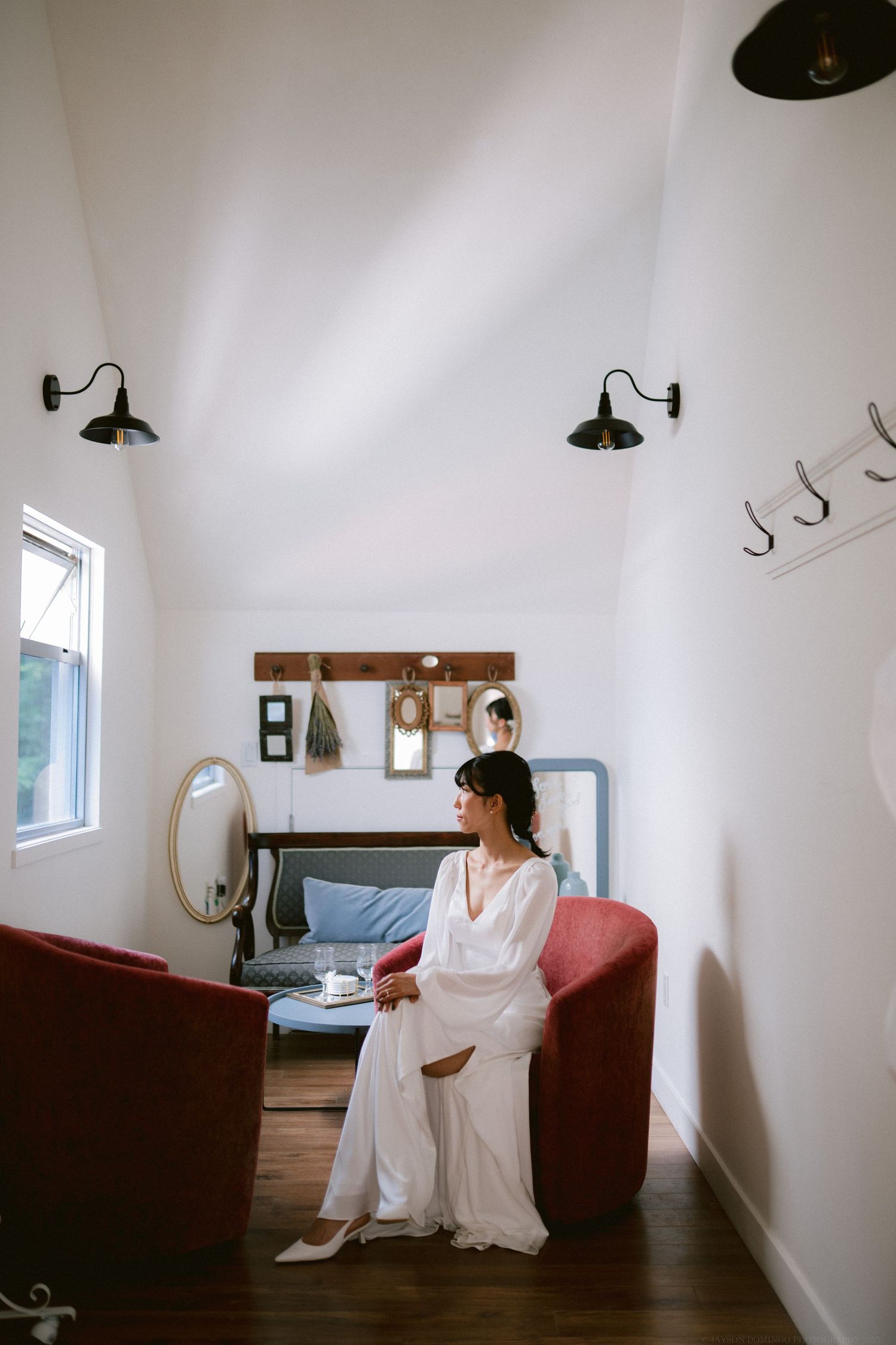 Interior room with white walls, vintage sconces, and a red velvet chair by a window.