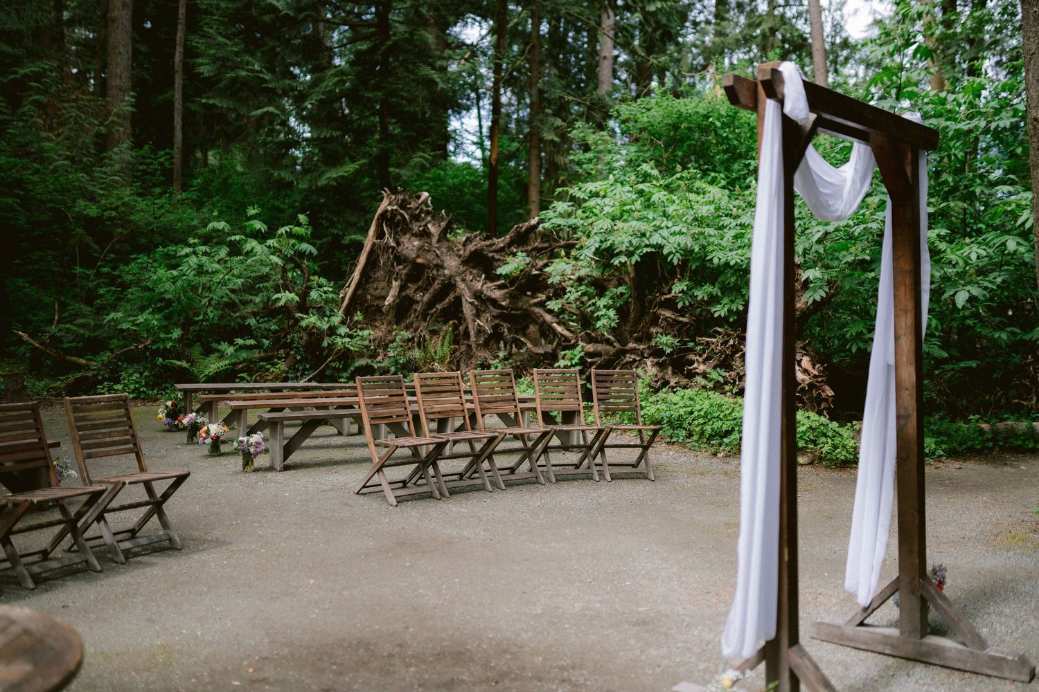 An outdoor wedding ceremony setup in a forest clearing with wooden chairs and a white-draped arch.