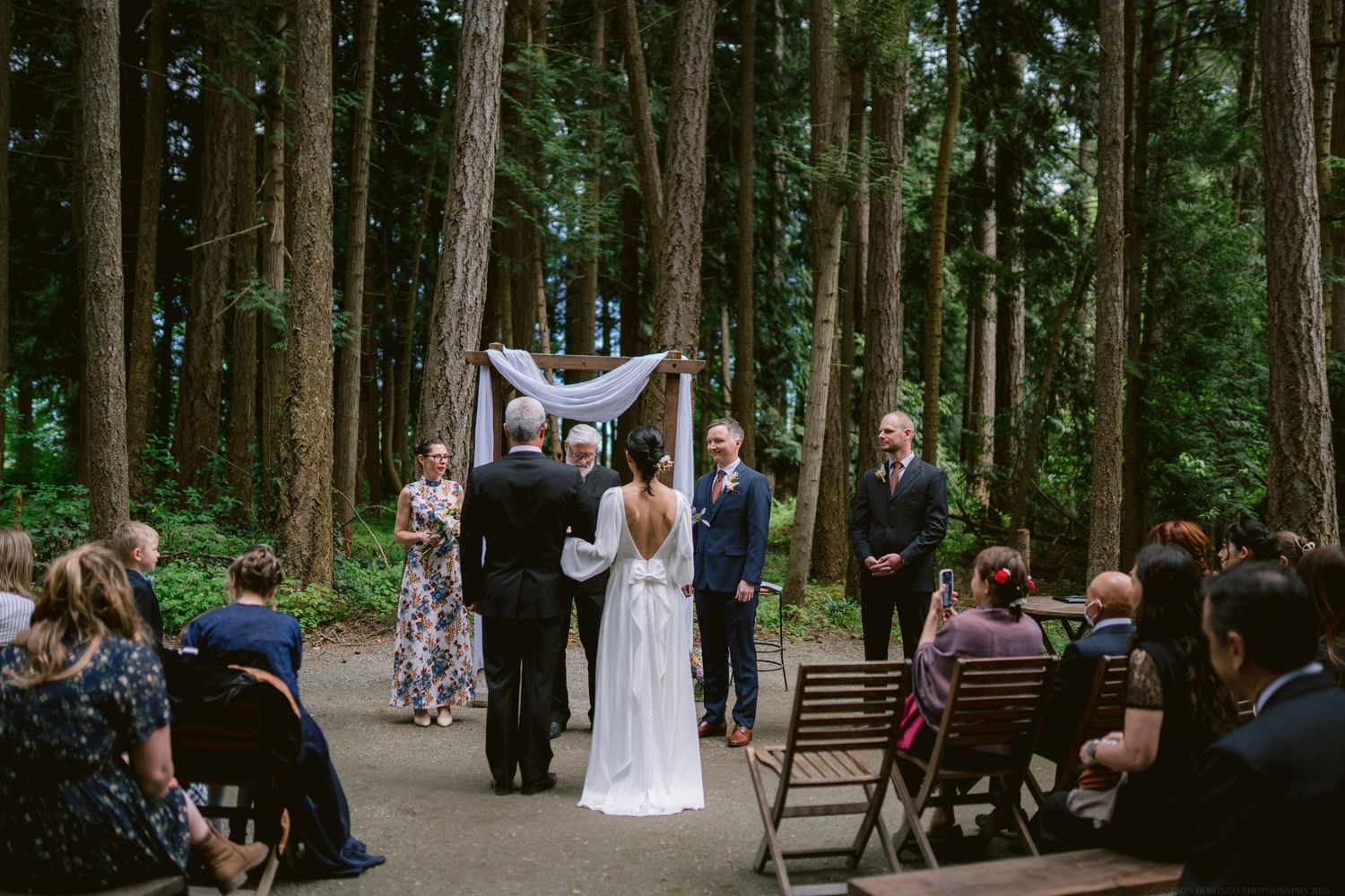 An intimate forest wedding ceremony with guests seated on wooden chairs among tall trees.