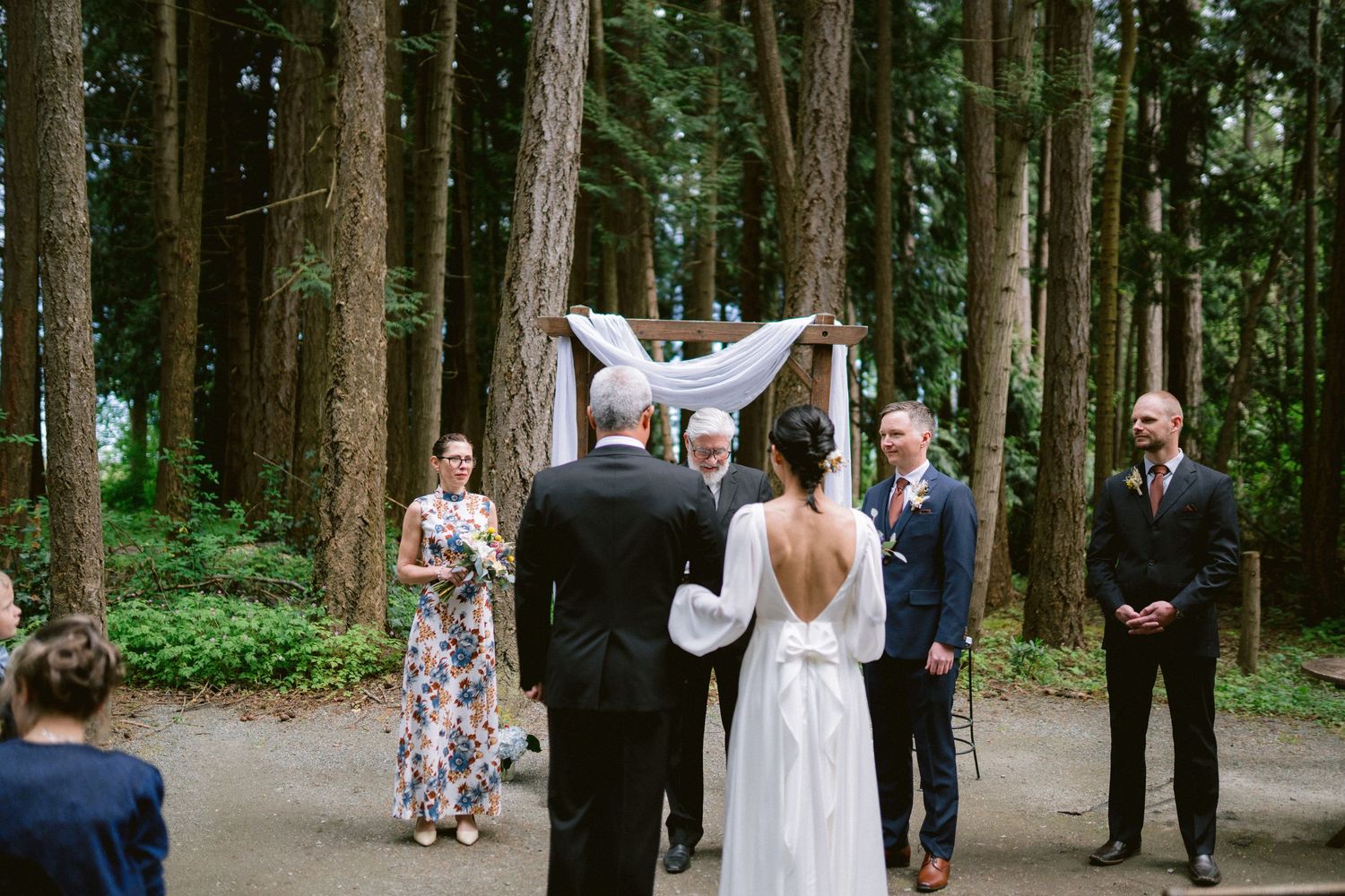 An outdoor forest wedding ceremony with a small group gathered around the bride and groom under tall trees.