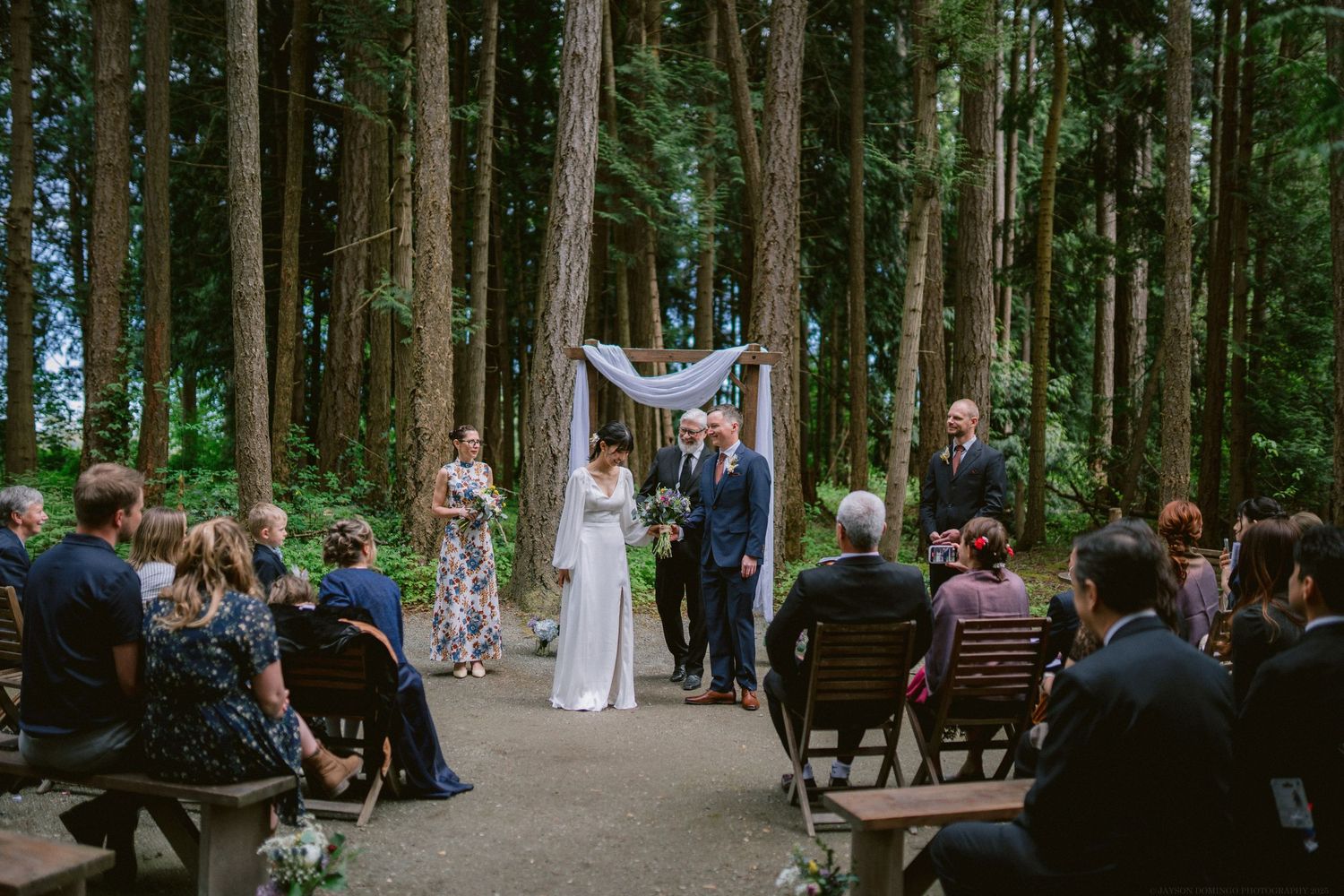A wedding ceremony takes place on a wooded path surrounded by tall evergreen trees as guests look on from wooden benches.