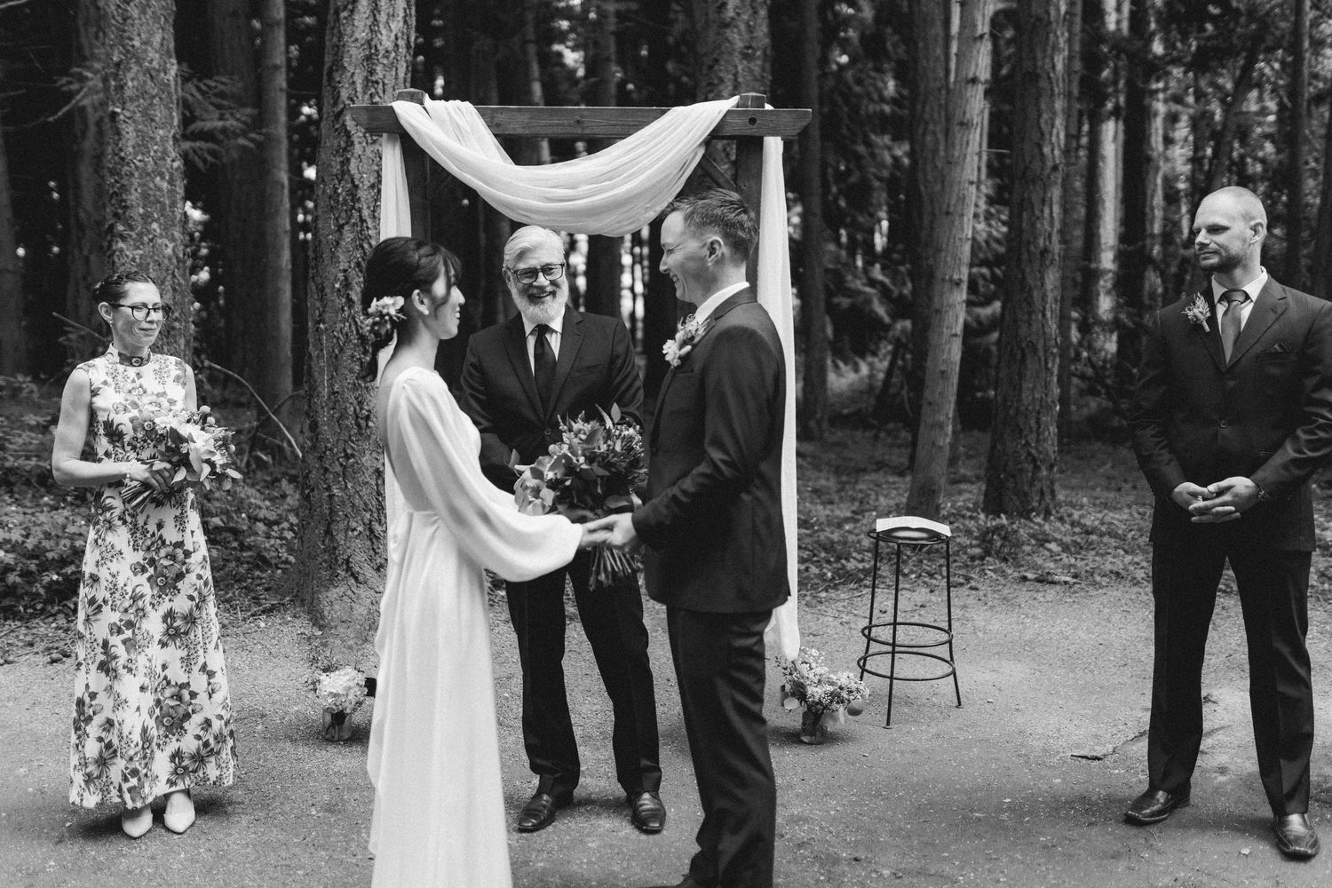 Black and white outdoor forest wedding ceremony with couple holding hands under white fabric canopy during intimate service.