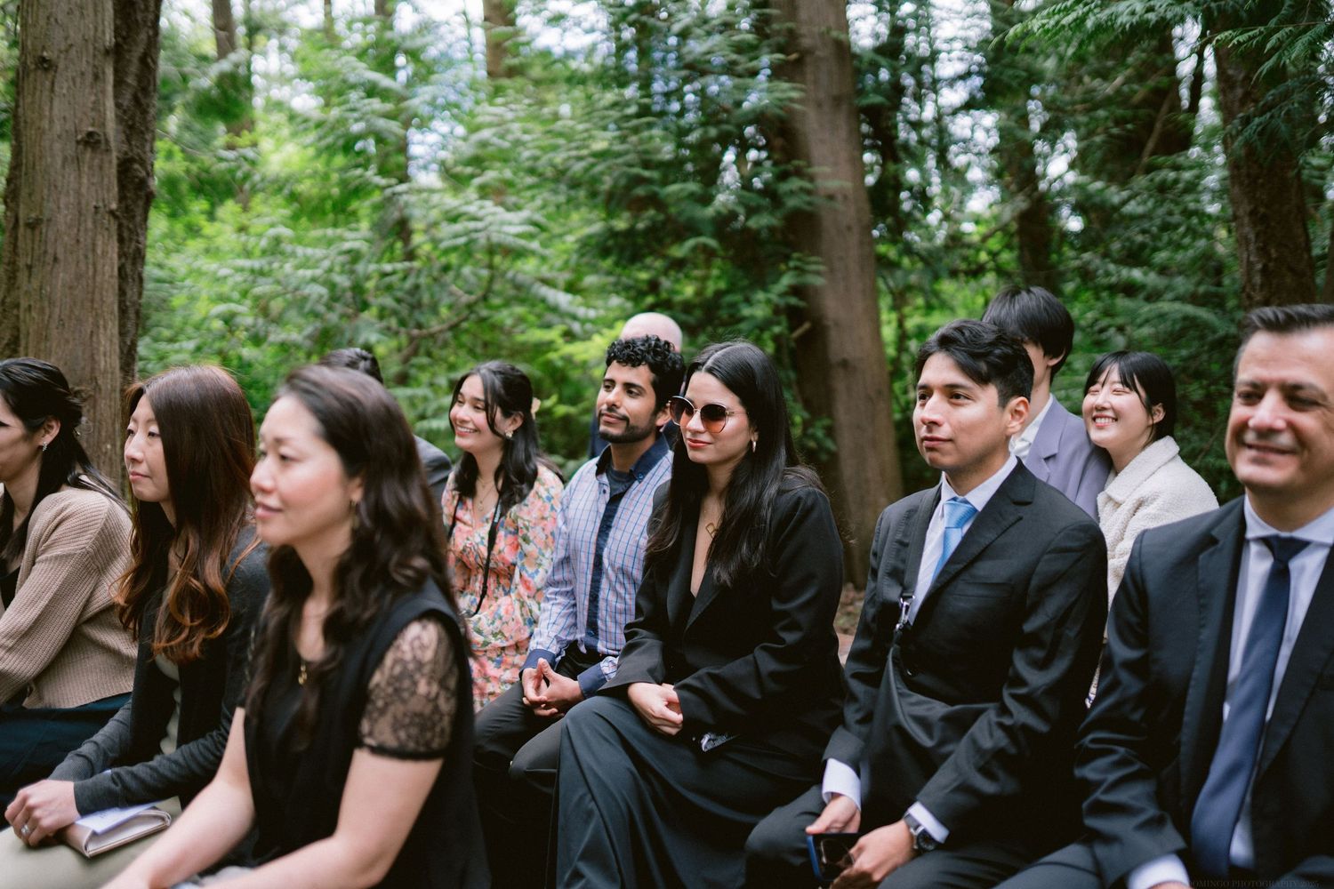 Wedding guests seated in a forest clearing smile and laugh during an outdoor ceremony.