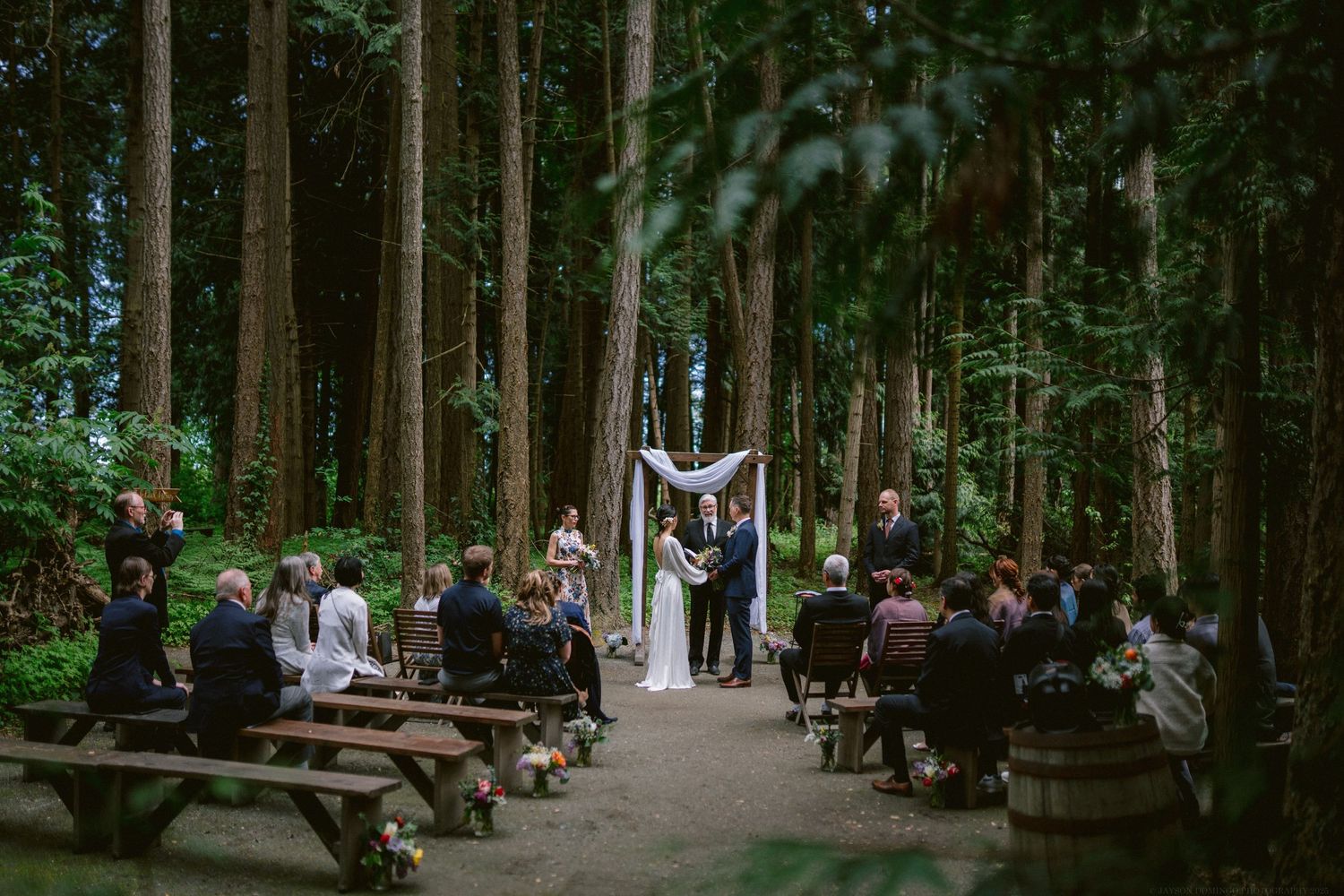 A wedding ceremony takes place on a path through tall evergreen trees with guests seated on wooden benches.