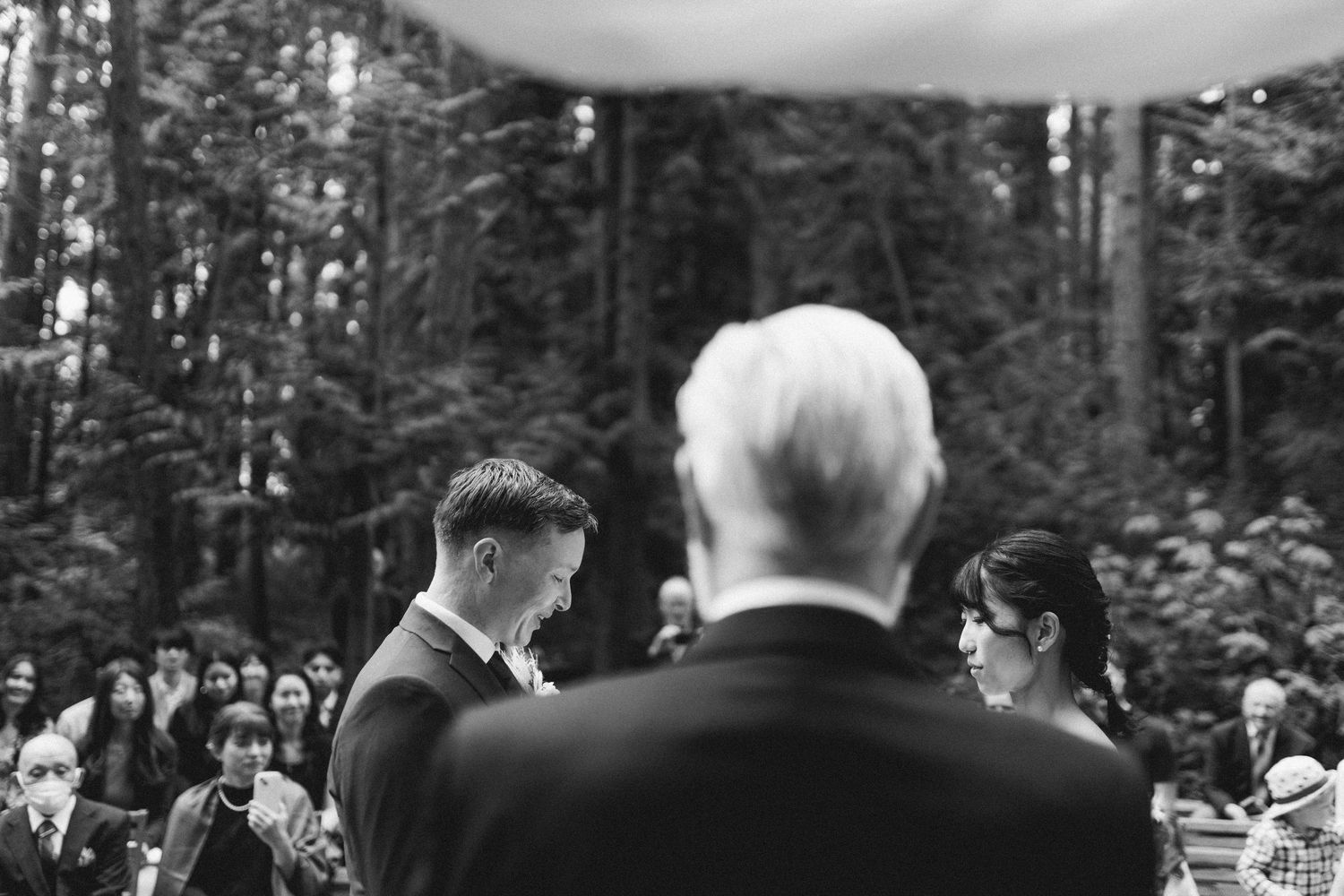 A black and white photo of a wedding ceremony in a forest with guests watching intently.