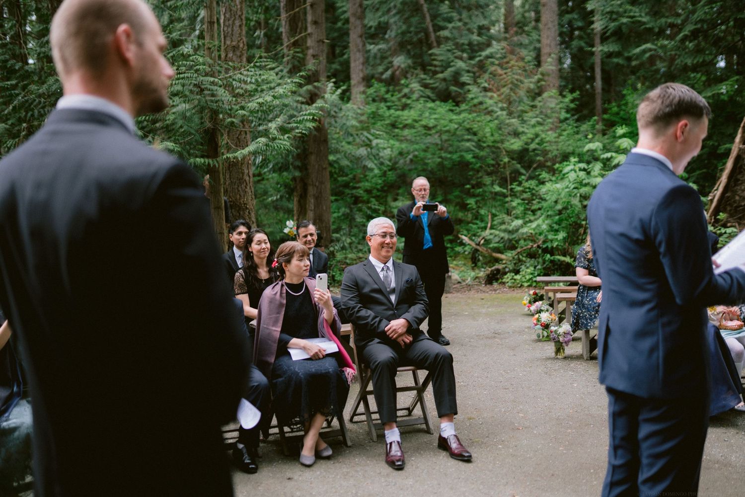 Wedding guests seated on wooden benches watch the outdoor forest ceremony.