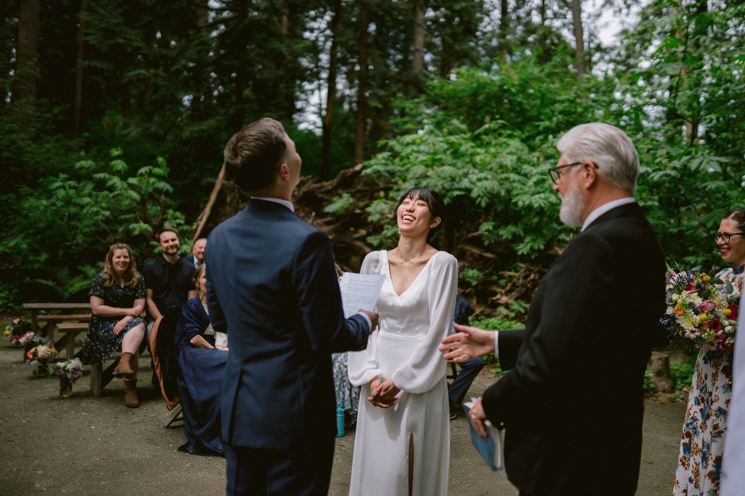 Outdoor wedding ceremony taking place in a lush forest setting with guests seated on wooden benches.