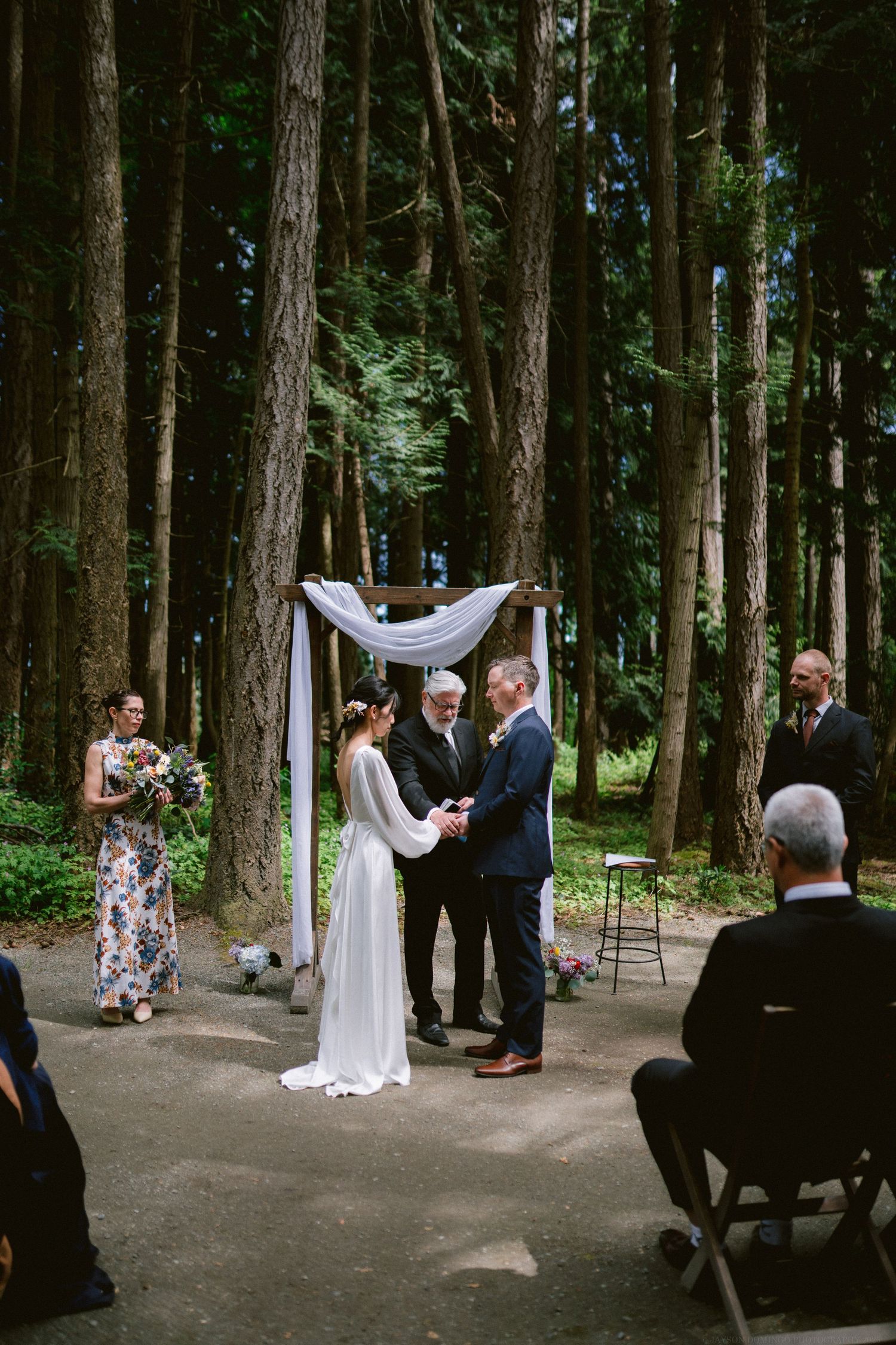 A wedding ceremony taking place on a path through tall trees with a white fabric arch and folding chairs.