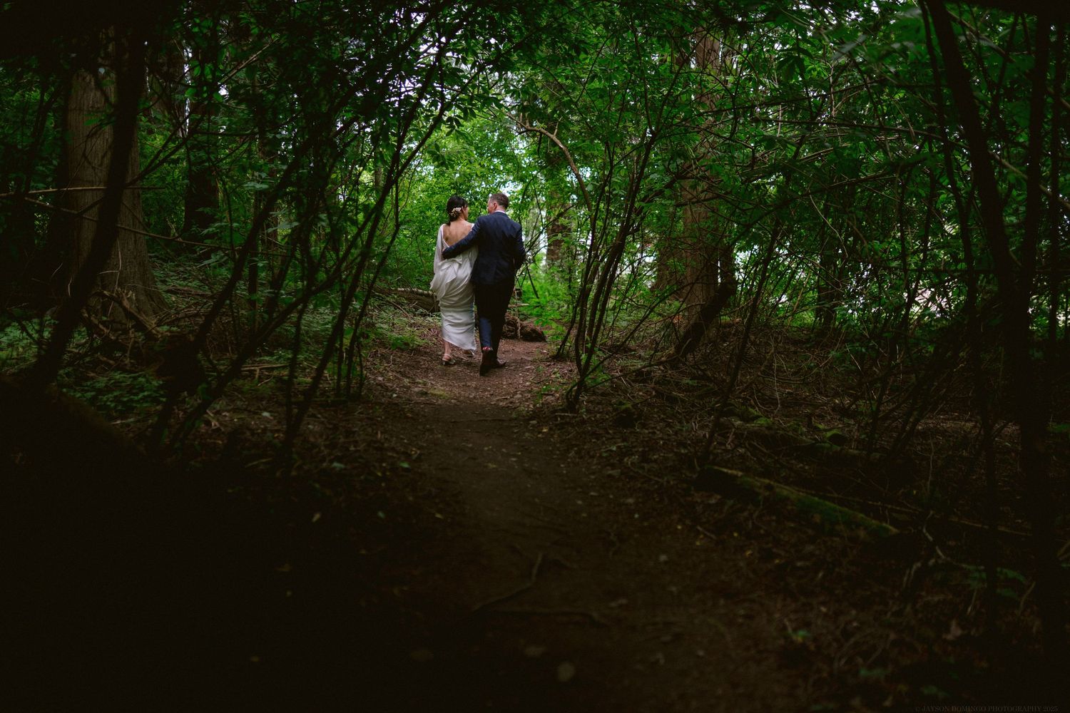 A silhouetted figure walks along a shaded forest path surrounded by lush green trees and foliage.