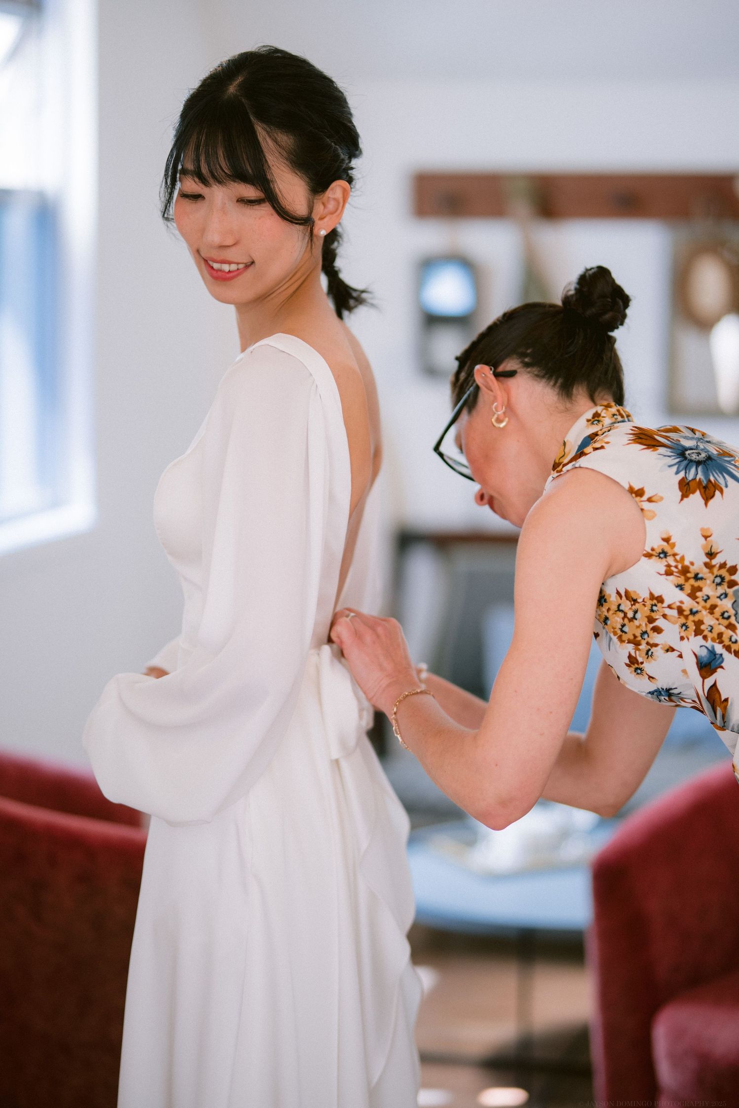 Someone helps adjust a flowing white dress during what appears to be a wedding preparation moment in a sunlit room.