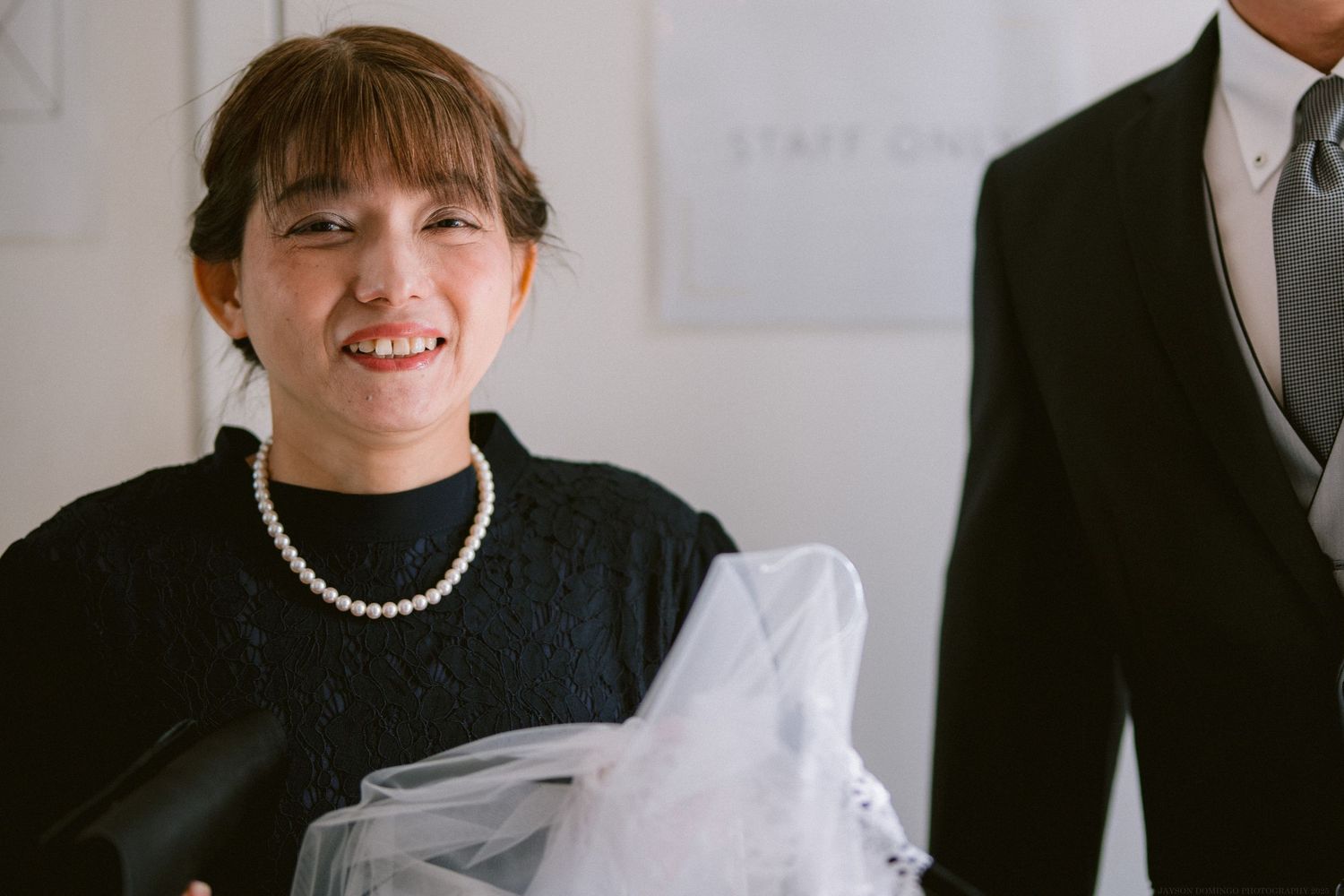 Wedding guest in black dress with pearl necklace smiling at formal event.