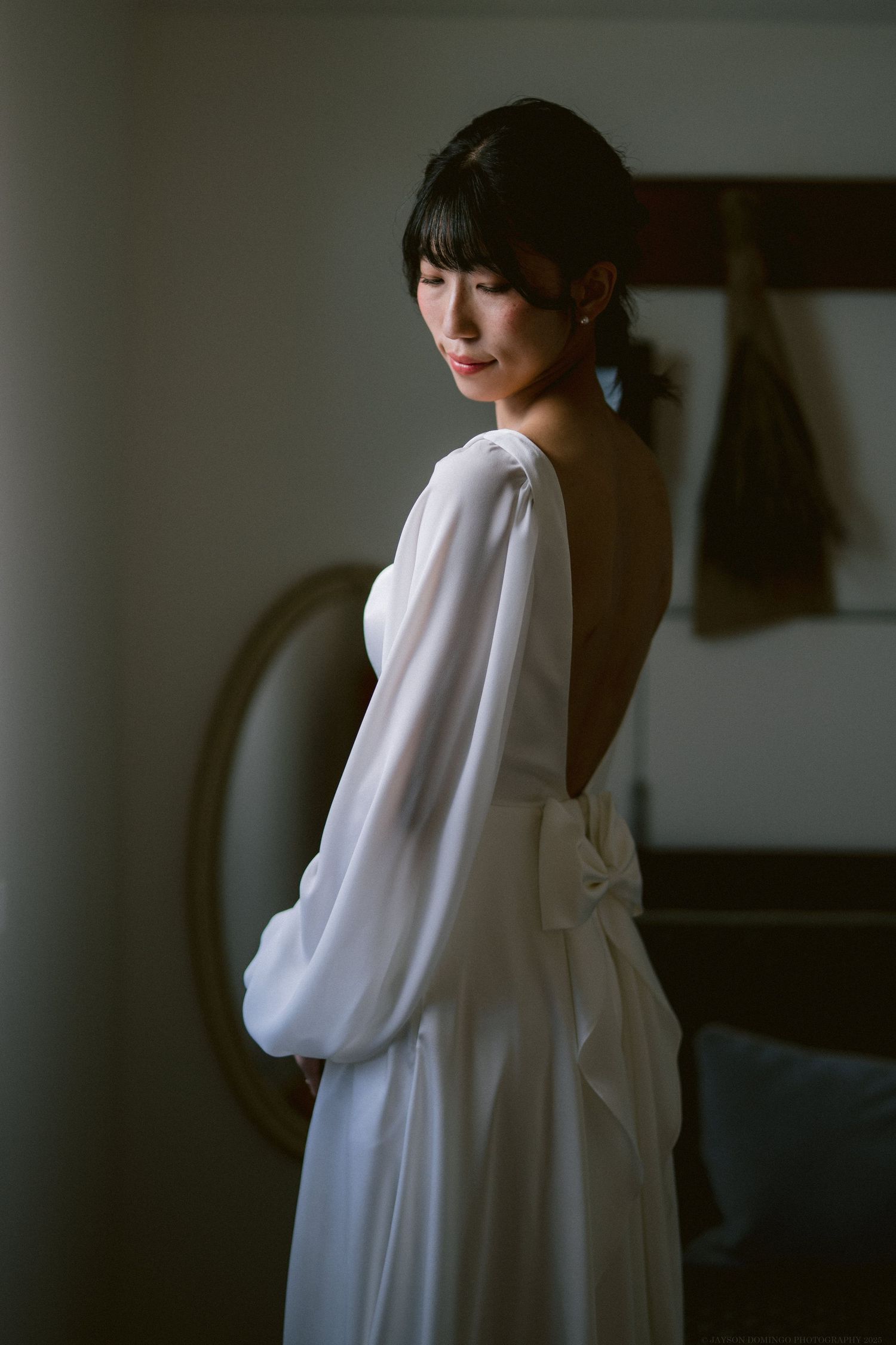 Moody portrait of bride in elegant white dress with low back and bow detail against dark background.