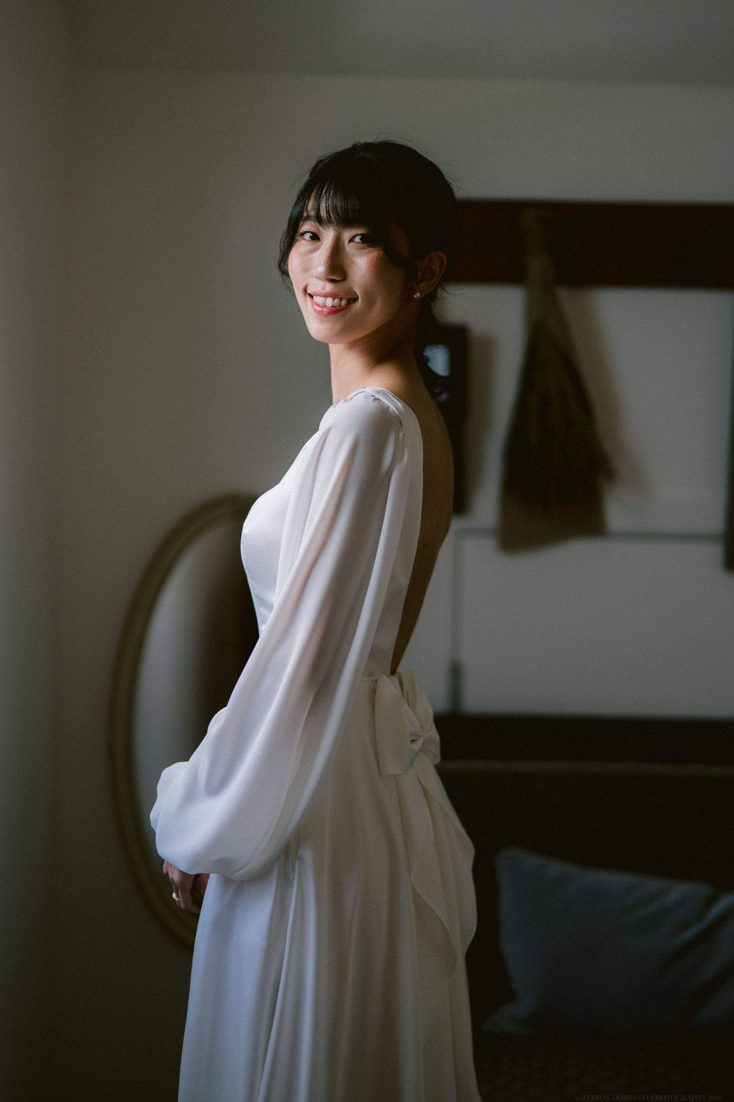 Side profile of bride in elegant white long-sleeve wedding dress against minimalist background.