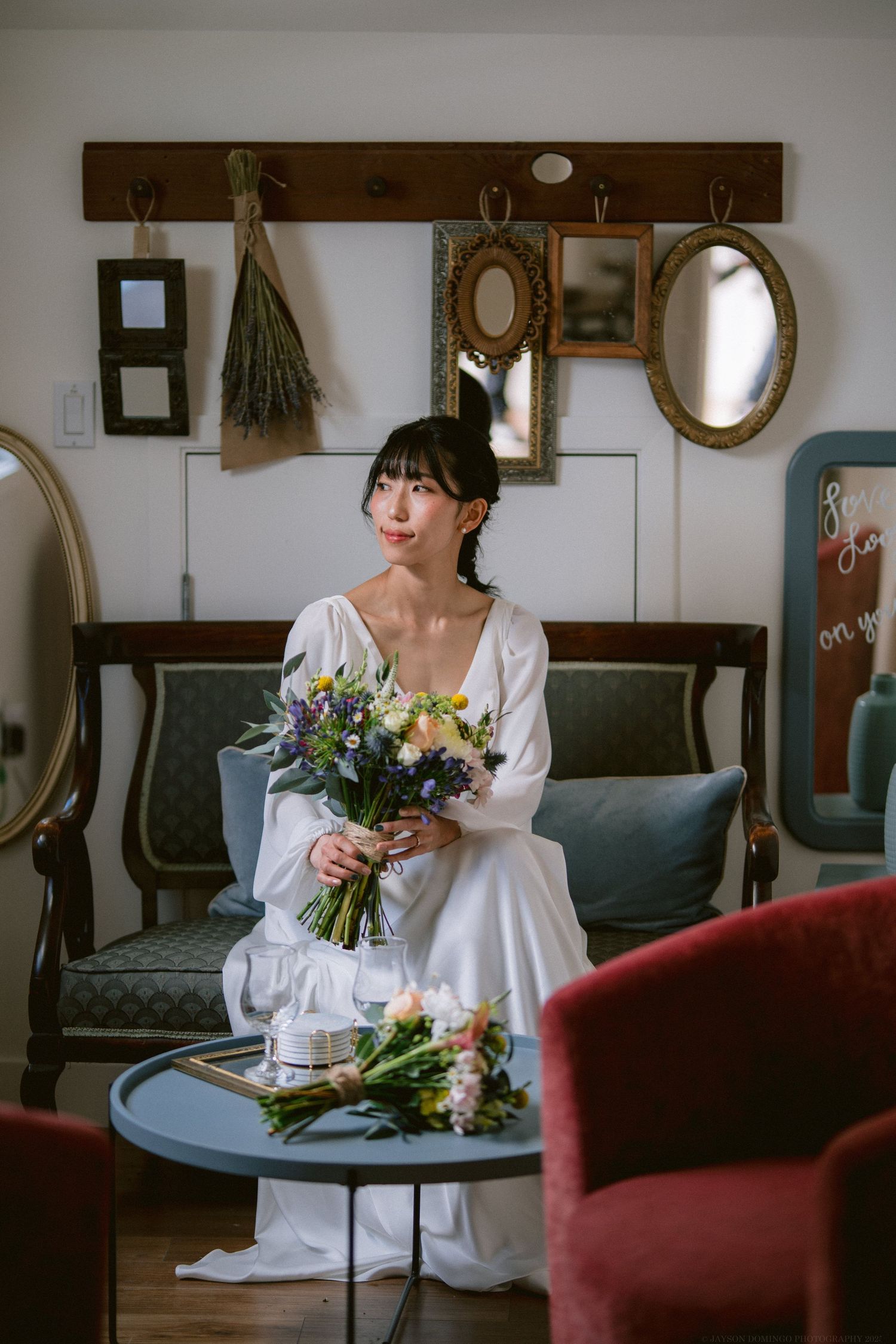 A white-dress figure sits on a vintage couch holding a fresh bouquet of flowers in a room with mirrors and wall hooks.
