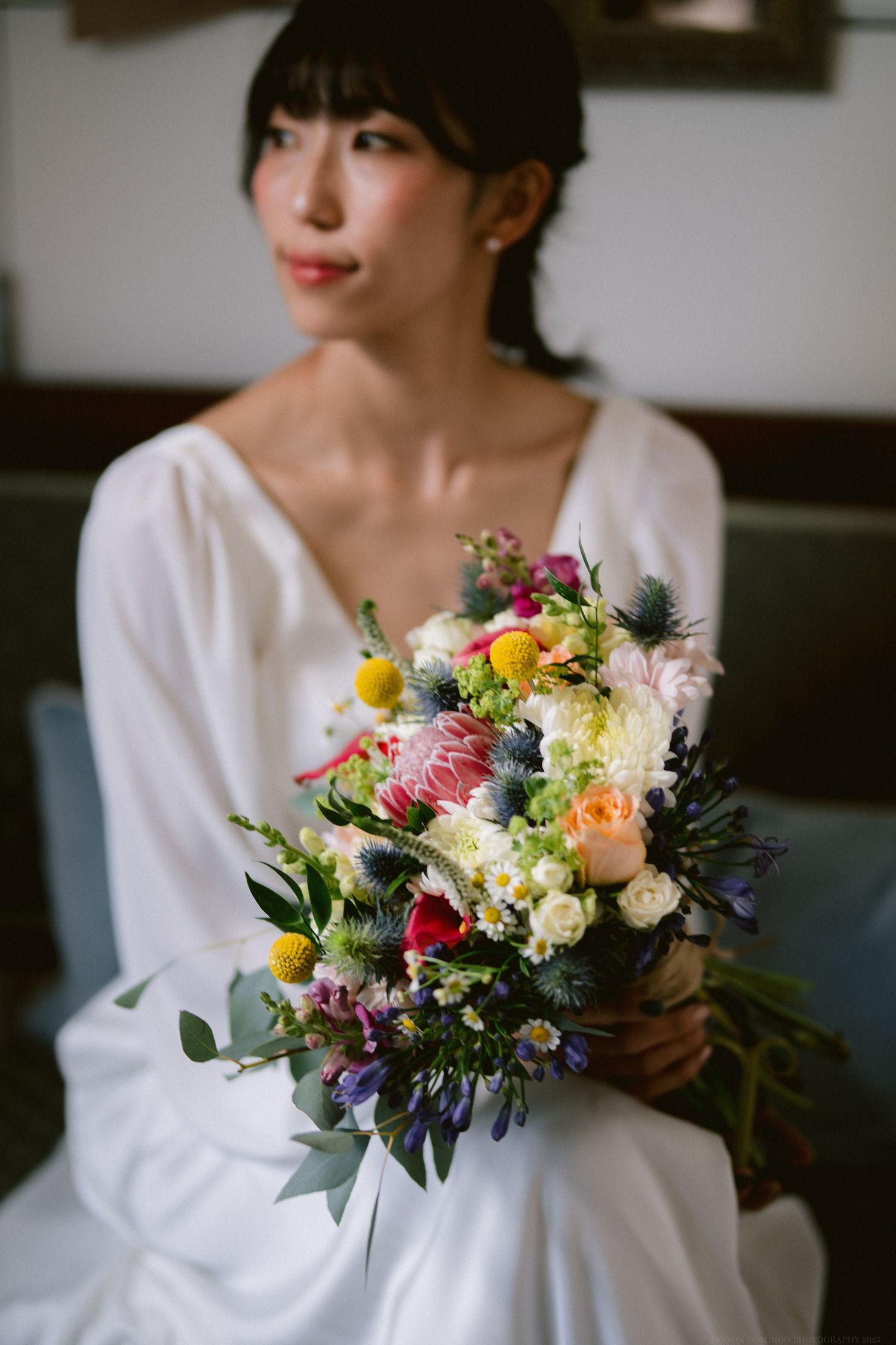 A colorful wildflower wedding bouquet held by someone wearing a white dress sitting indoors.