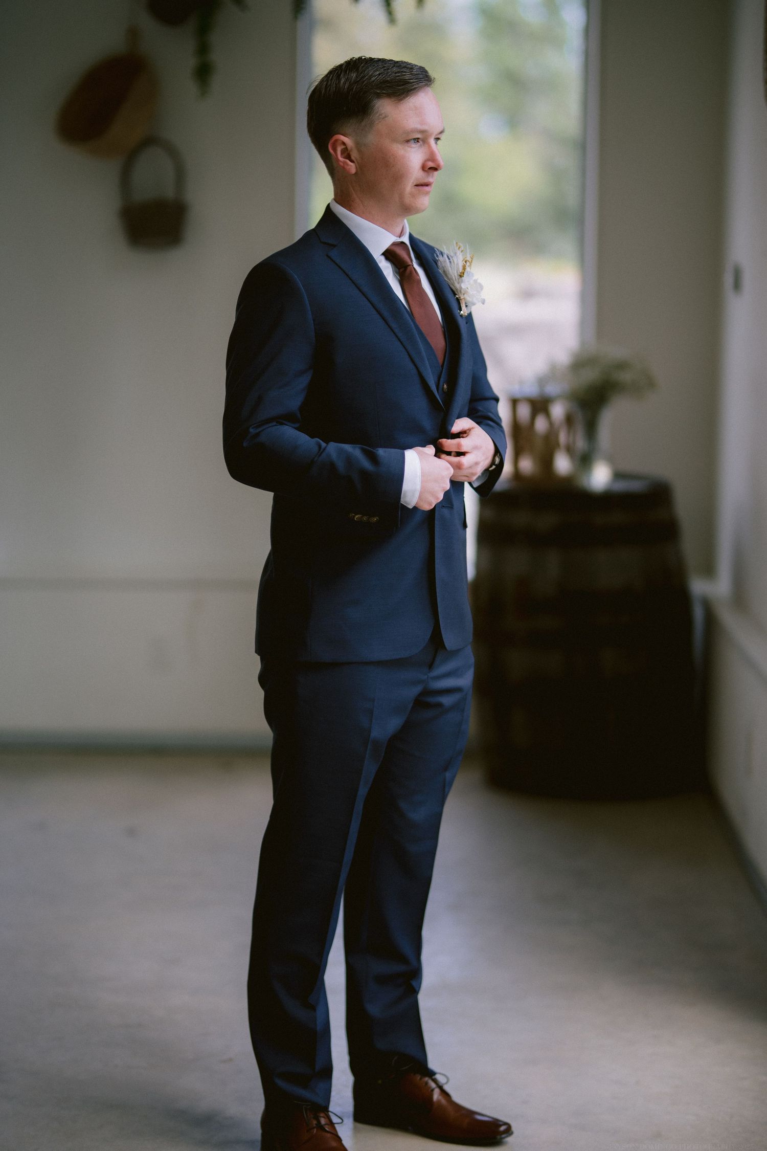 A groom in a navy blue suit and brown shoes stands by a rustic barrel in an elegant venue.