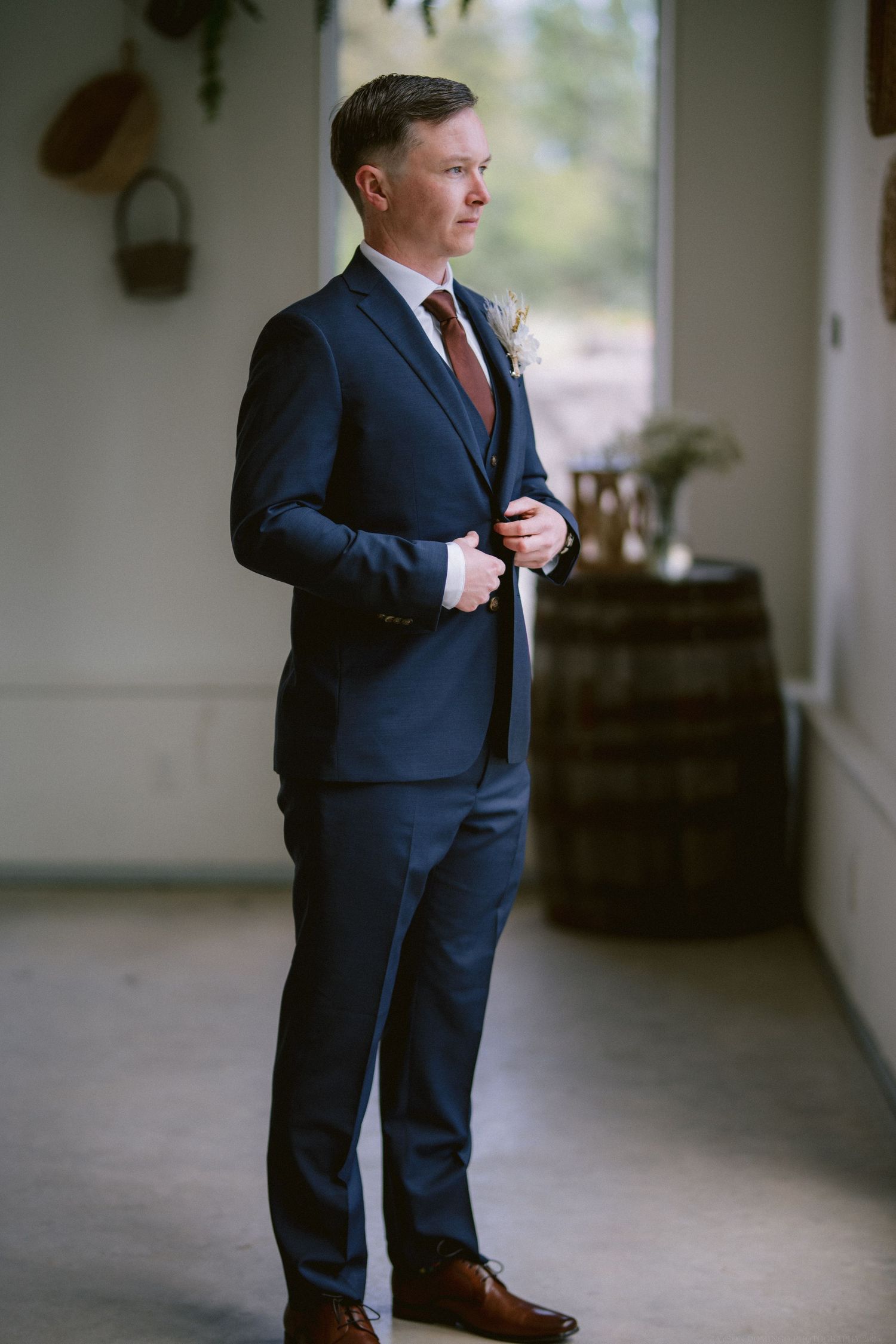 A groom in a navy blue suit and brown leather shoes stands thoughtfully by a wooden barrel in a rustic venue.