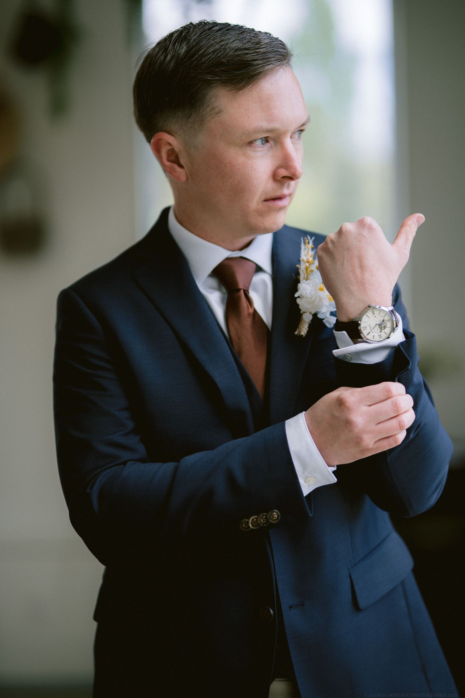 A groom in a navy suit and brown tie adjusts his cuff while wearing a boutonniere and wristwatch.