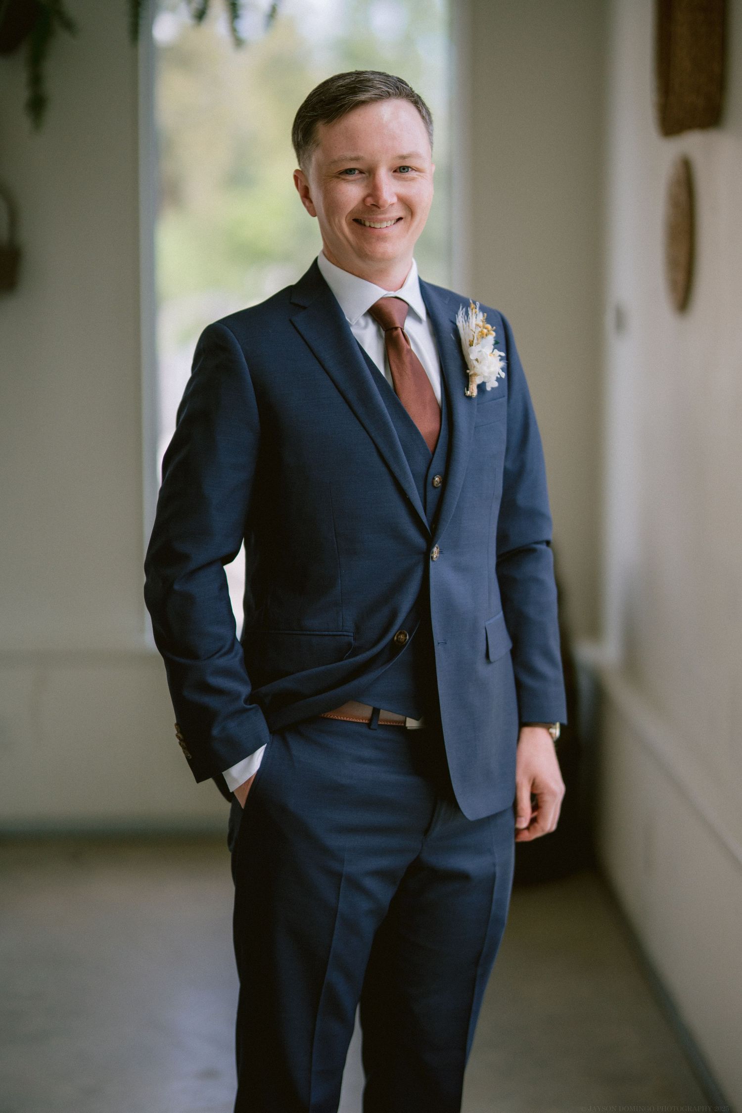 Groom in navy blue three-piece suit with rust-colored tie and boutonniere poses in sunlit hallway.