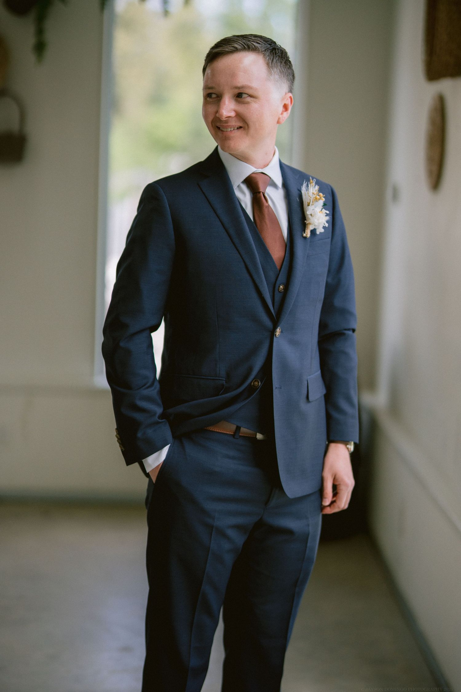 A groom in a navy blue suit with brown tie and boutonniere stands in a dim hallway, smiling warmly.