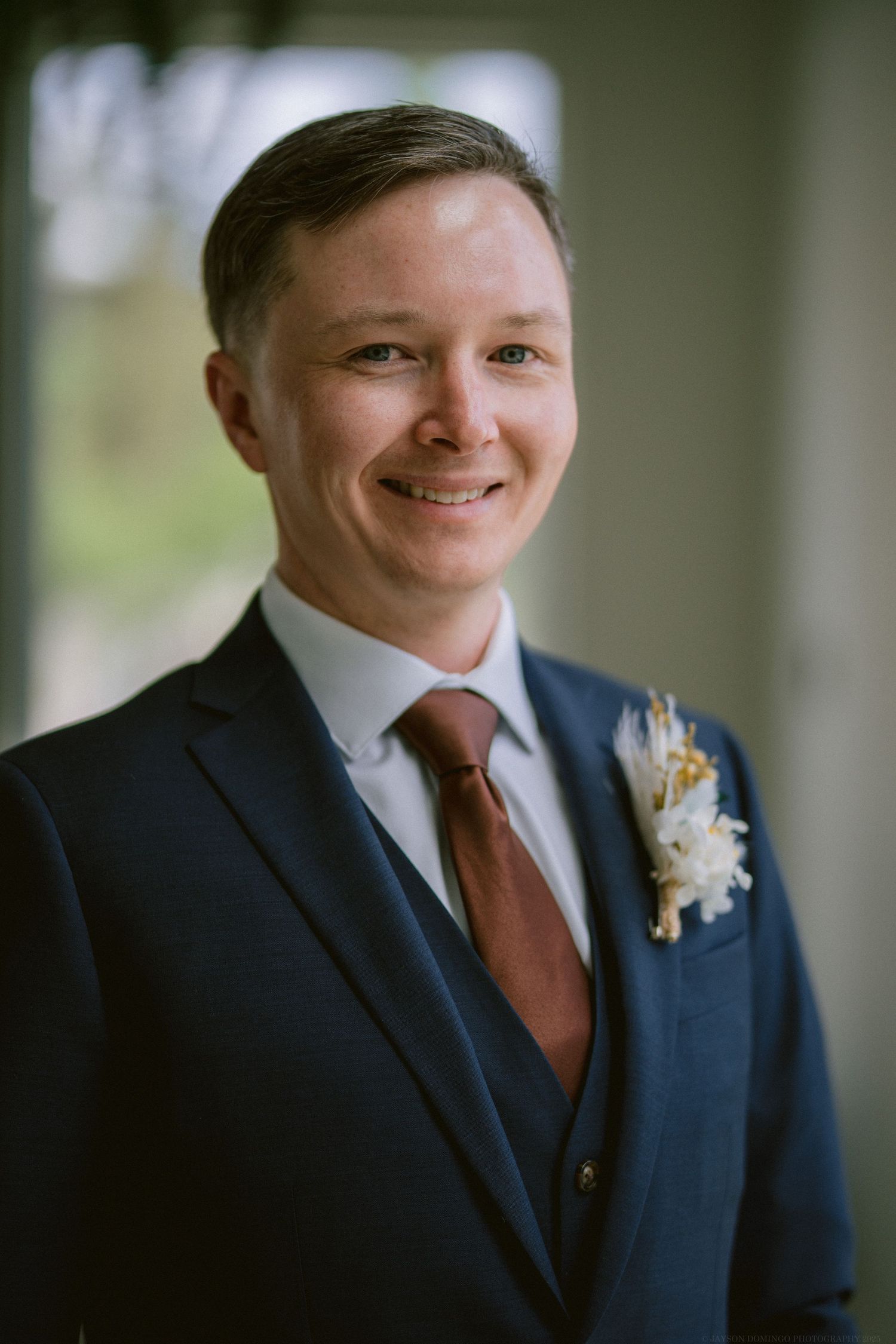 A groom wearing a navy blue suit with burgundy tie and boutonniere smiles warmly at the camera.