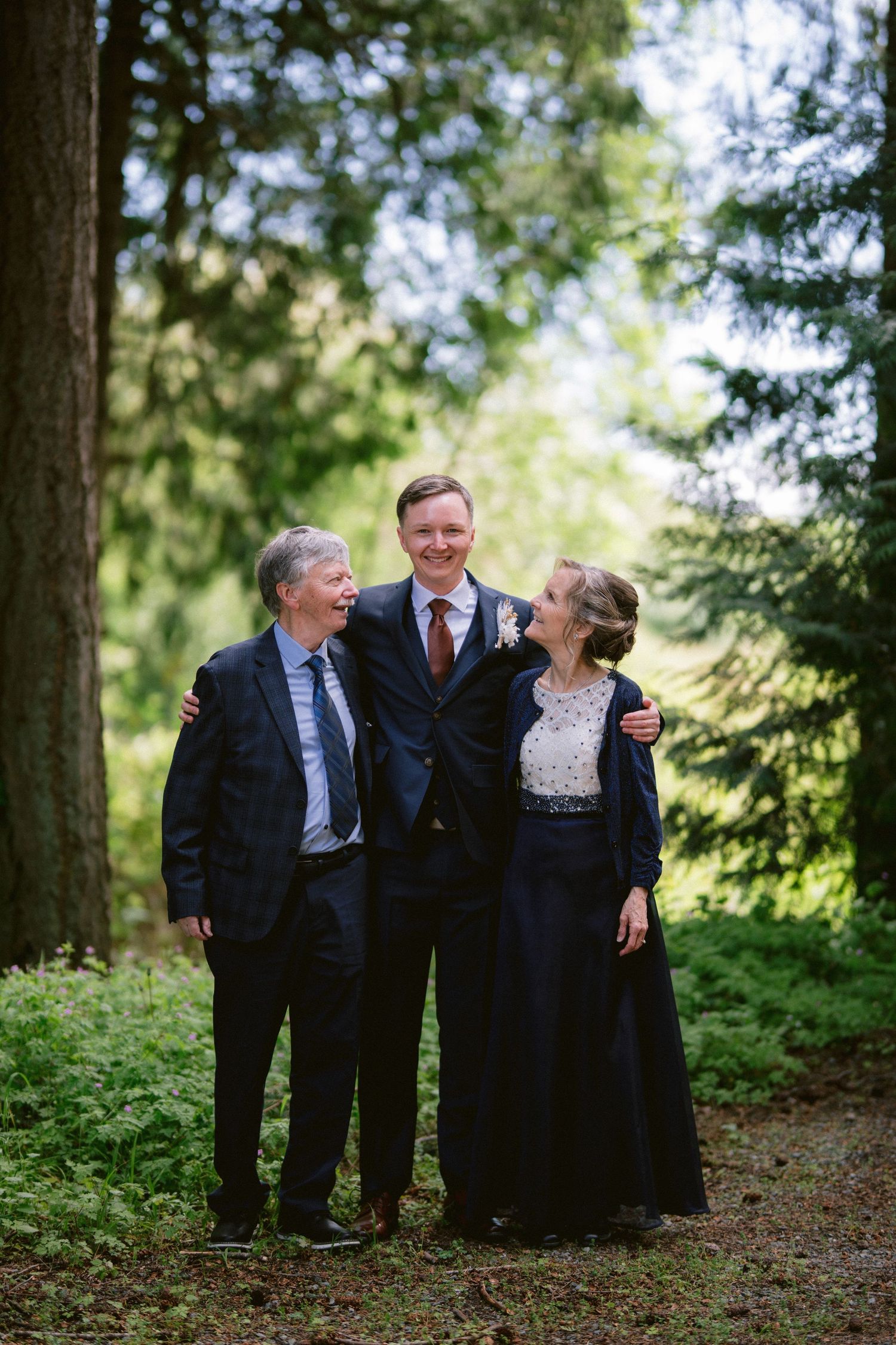 Three people in formal attire share a joyful moment together on a wooded path surrounded by evergreen trees.