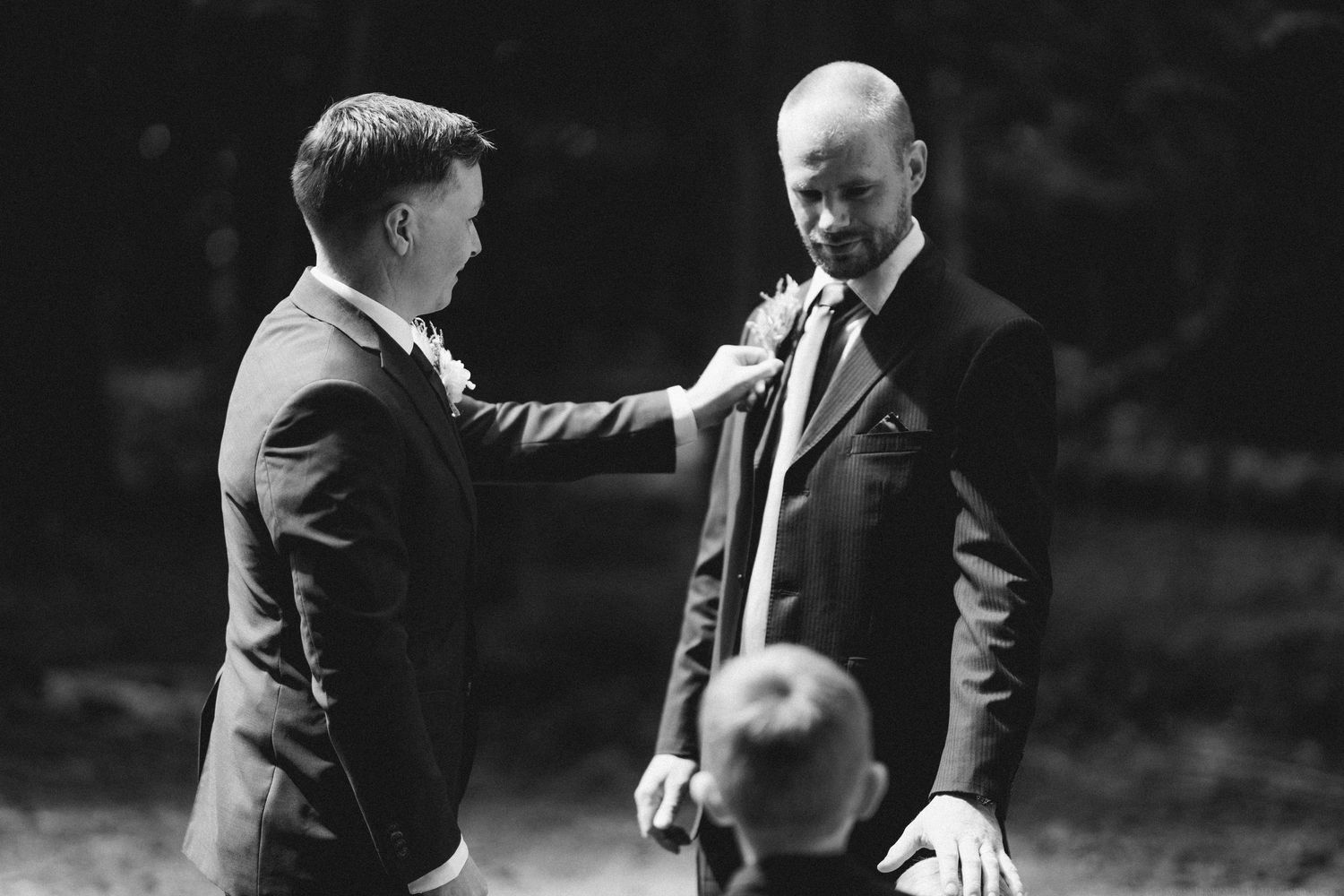 Black and white photo of groom being helped with his tie by another person during wedding preparations outdoors.