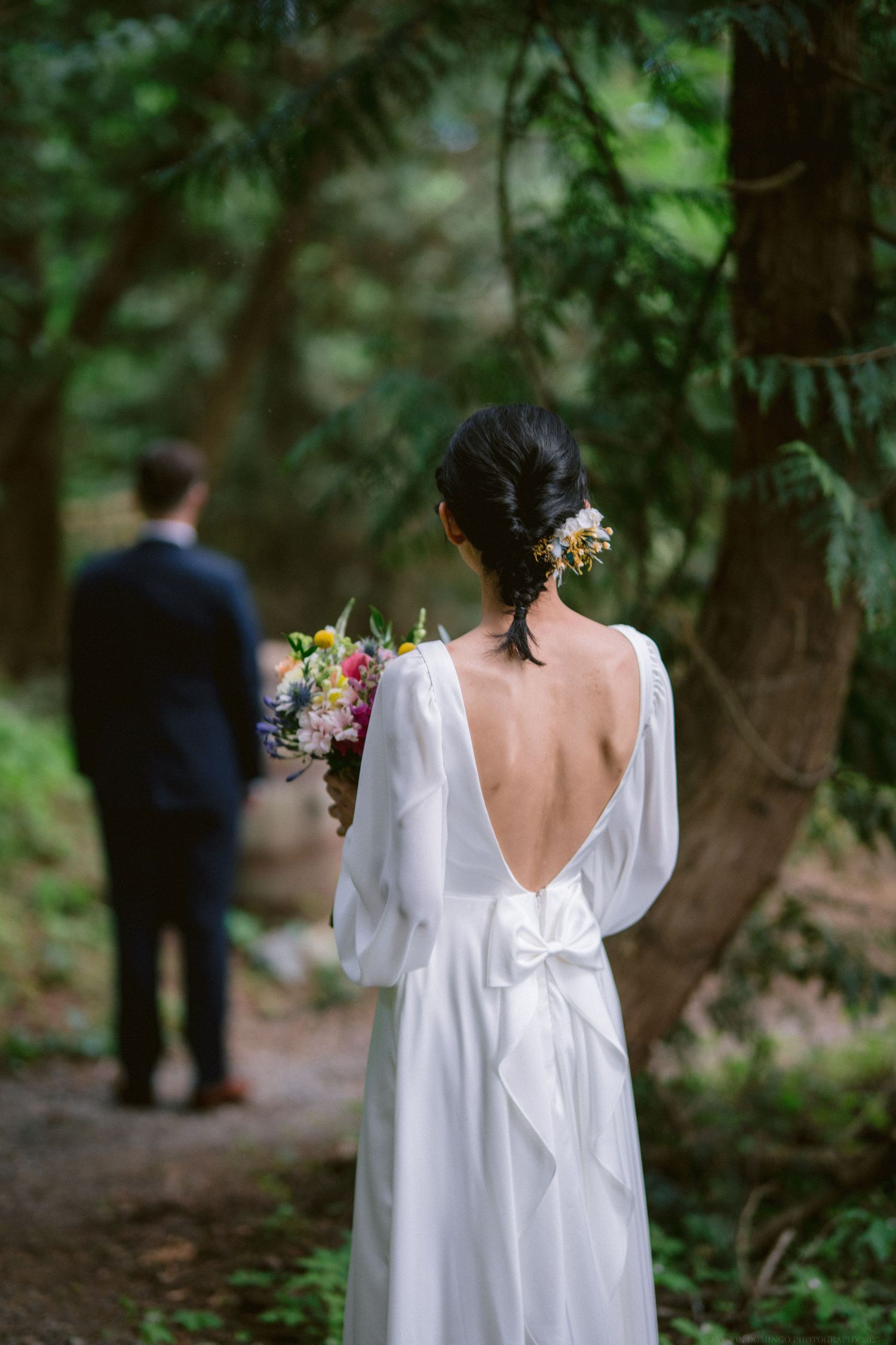 A bride in a backless white wedding dress holds a bouquet while looking towards someone in a wooded setting.