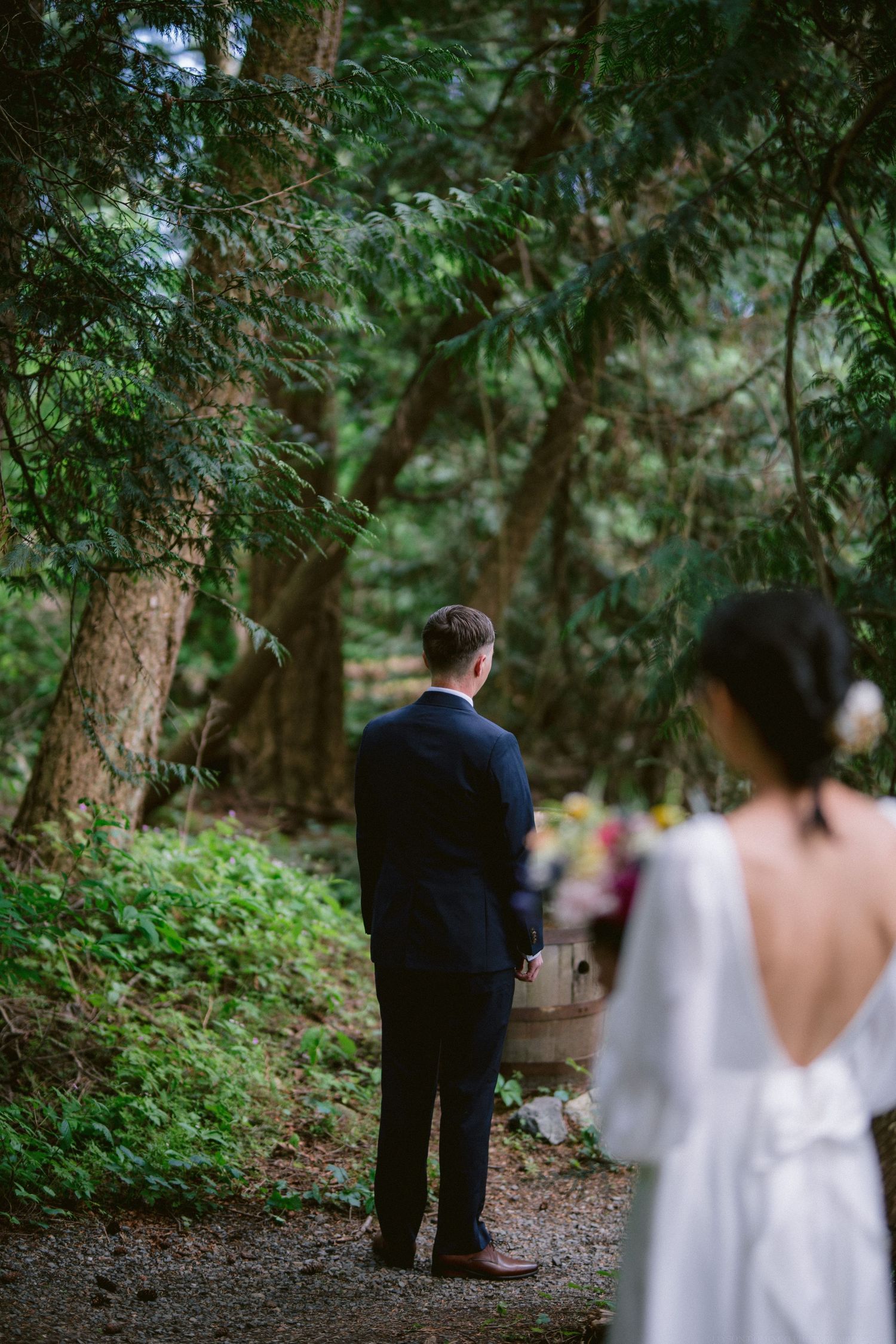 A couple shares an intimate first look moment on a forest path surrounded by lush greenery and tall trees.