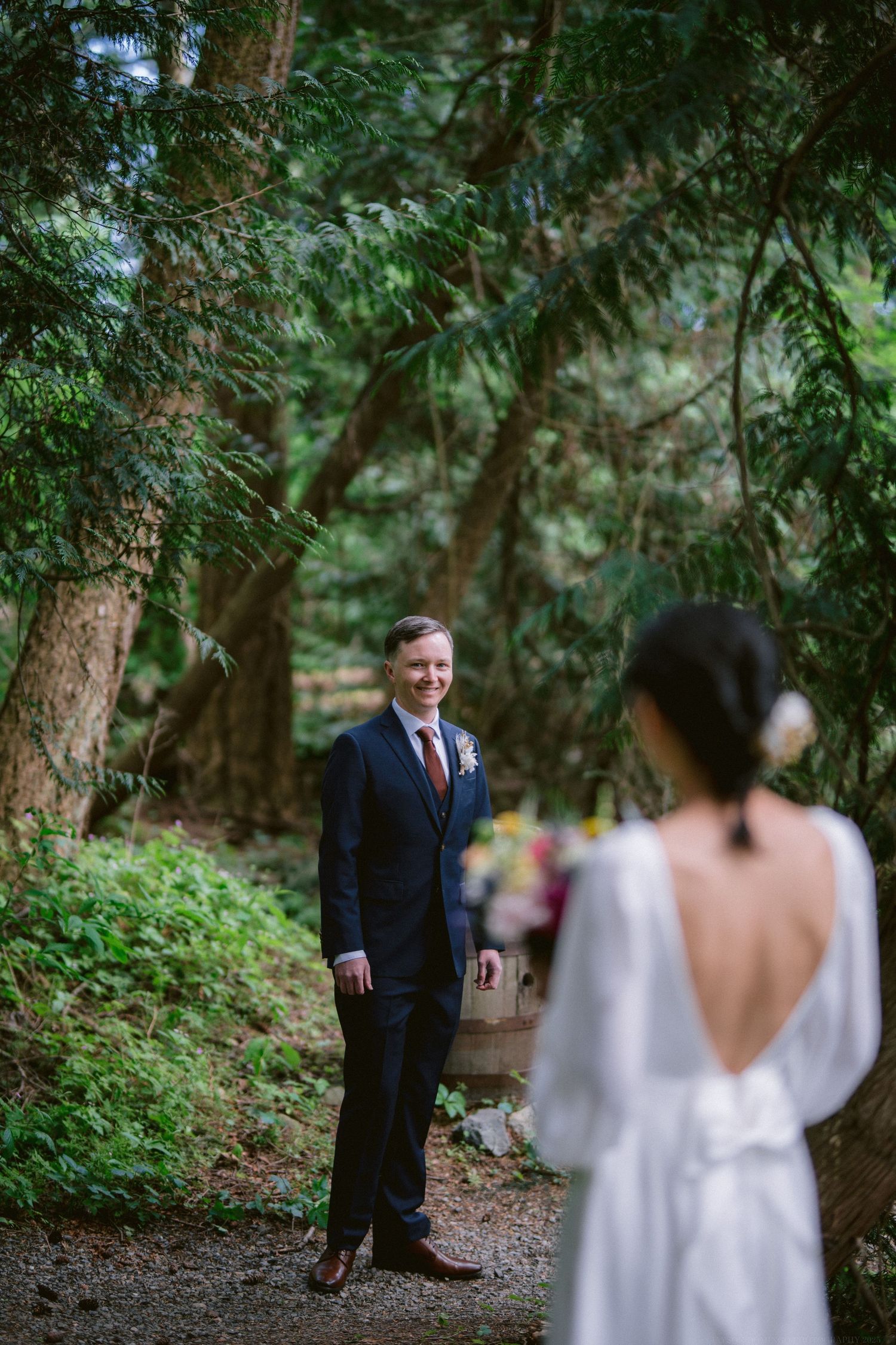 First look wedding photo of groom waiting on forest path as bride in white dress approaches from behind.