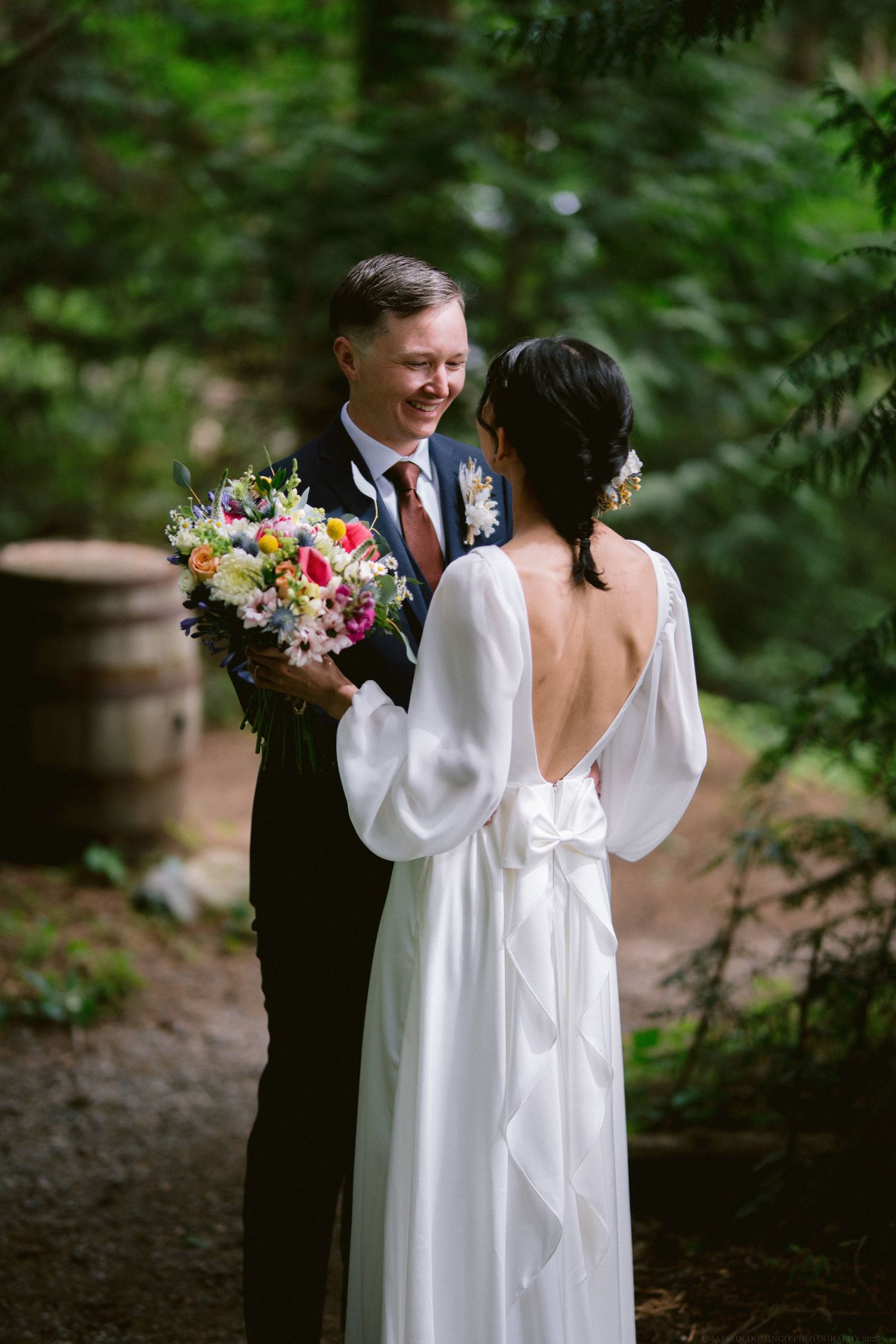 A wedding couple shares a tender moment in a wooded outdoor setting with a colorful bouquet of flowers.