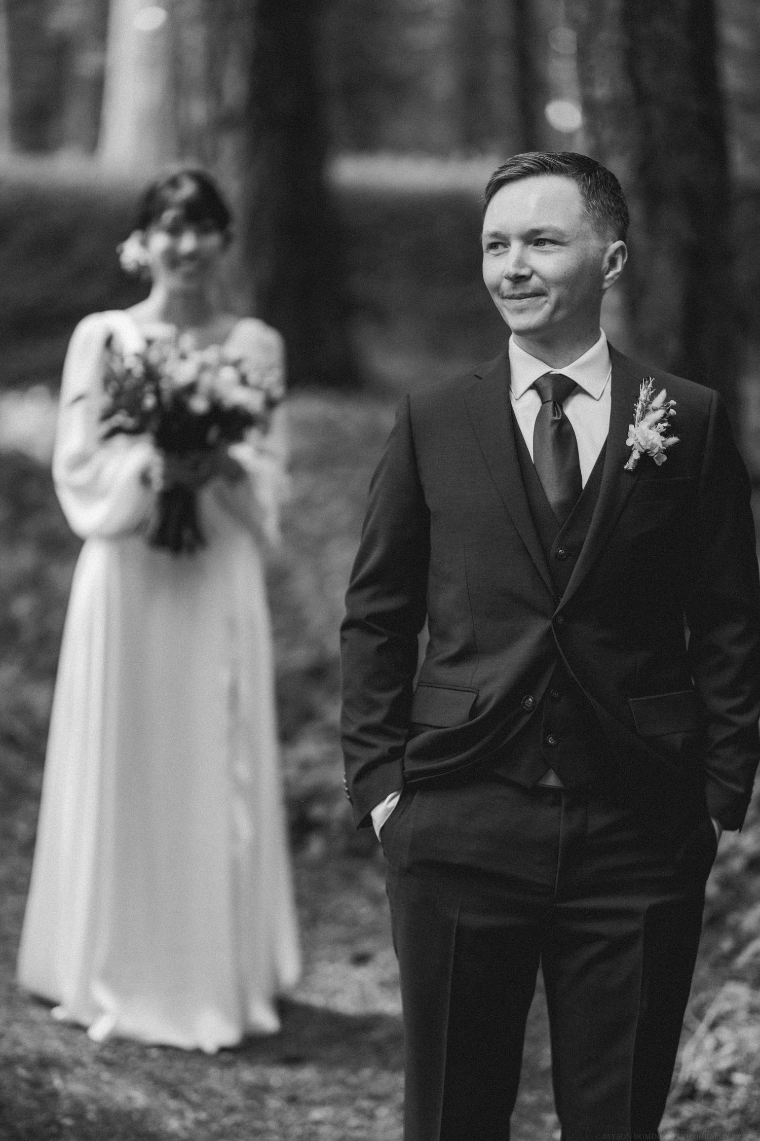 Black and white wedding photo of bride holding bouquet in background while groom smiles in foreground in forest setting.